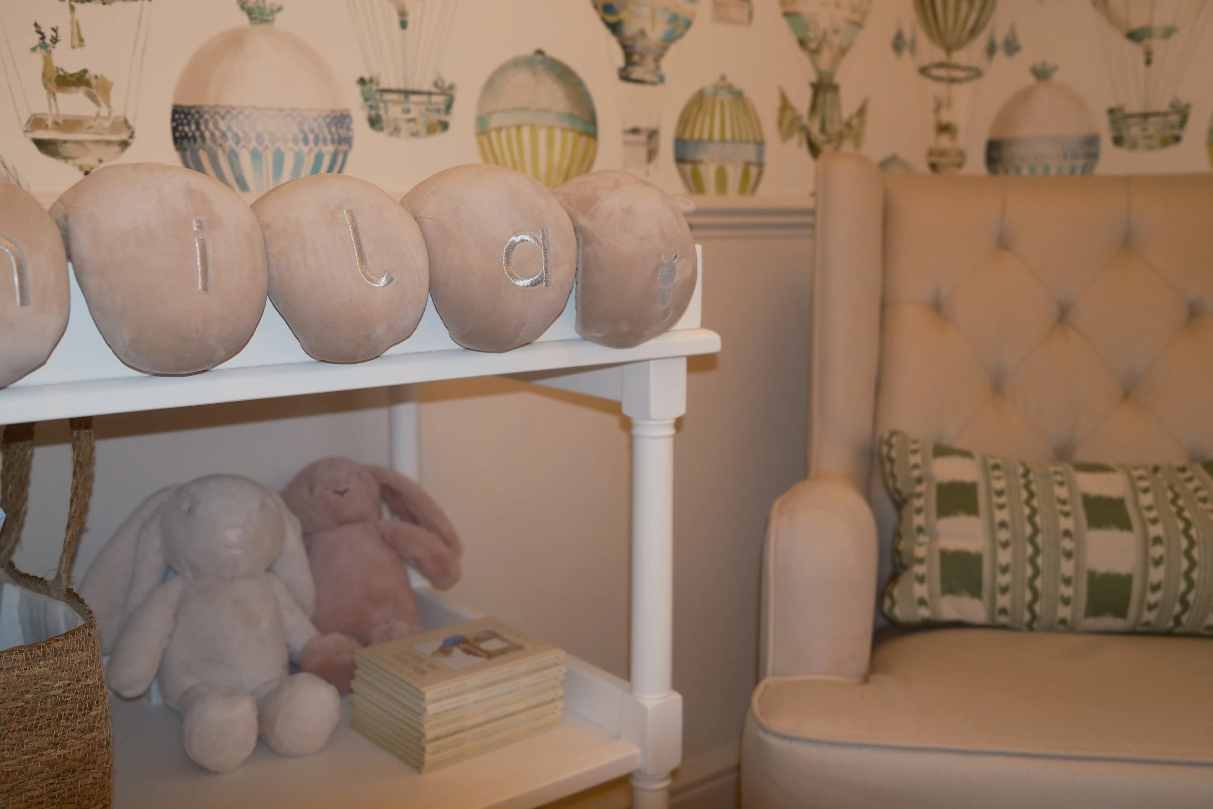 A close up image of some bunnies and books on a changing table shelf next to a cream rocking chair with designer green patterned cushion and hot air balloon wallpaper