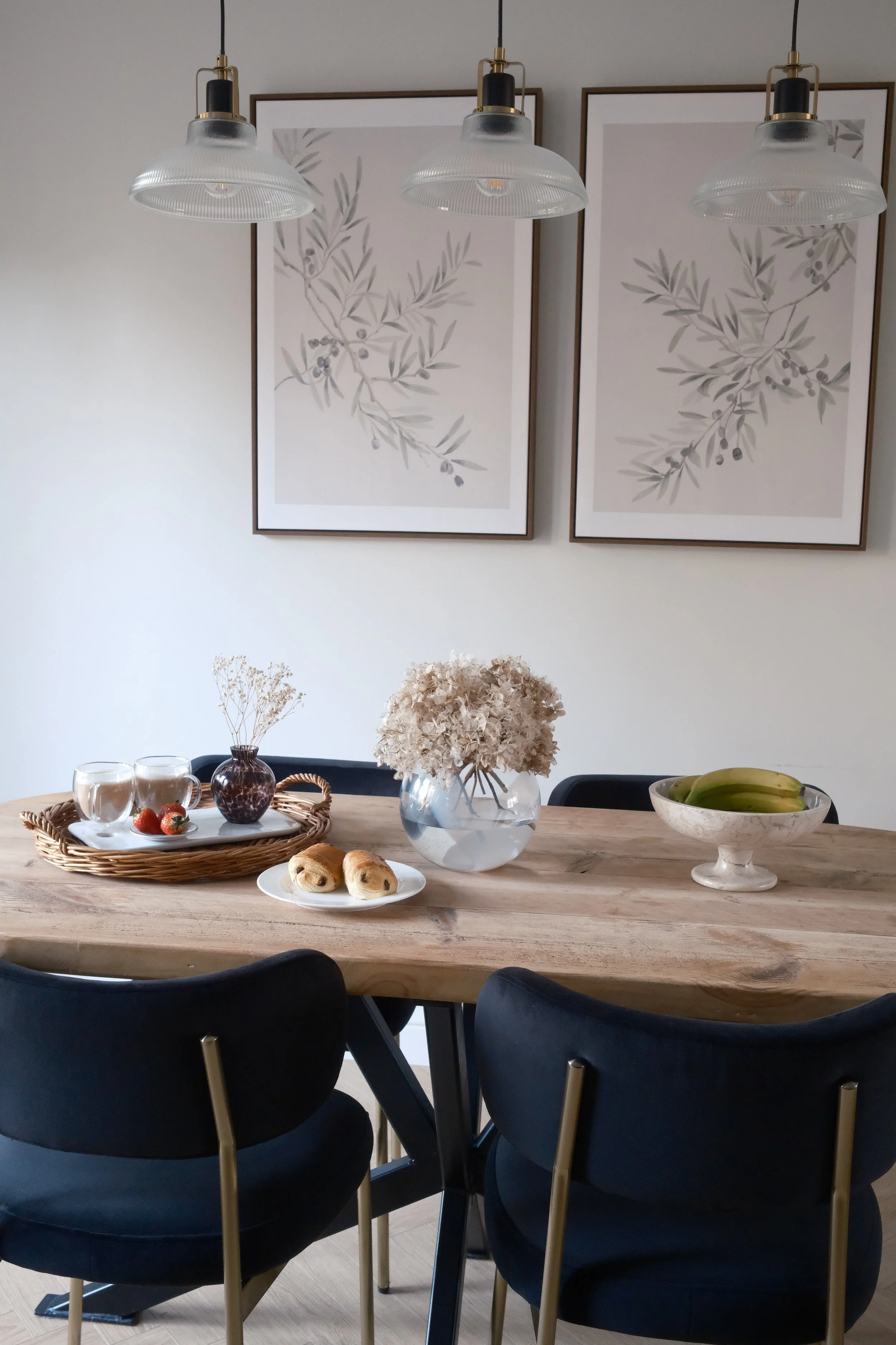A modern dining room with a wooden table, black chairs with gold legs, and decor including a vase of dried flowers, a bowl of bananas, a plate of pastries, and a tray with glasses and strawberries. There are two framed botanical prints on the white wall and three hanging pendant lights above the table.
