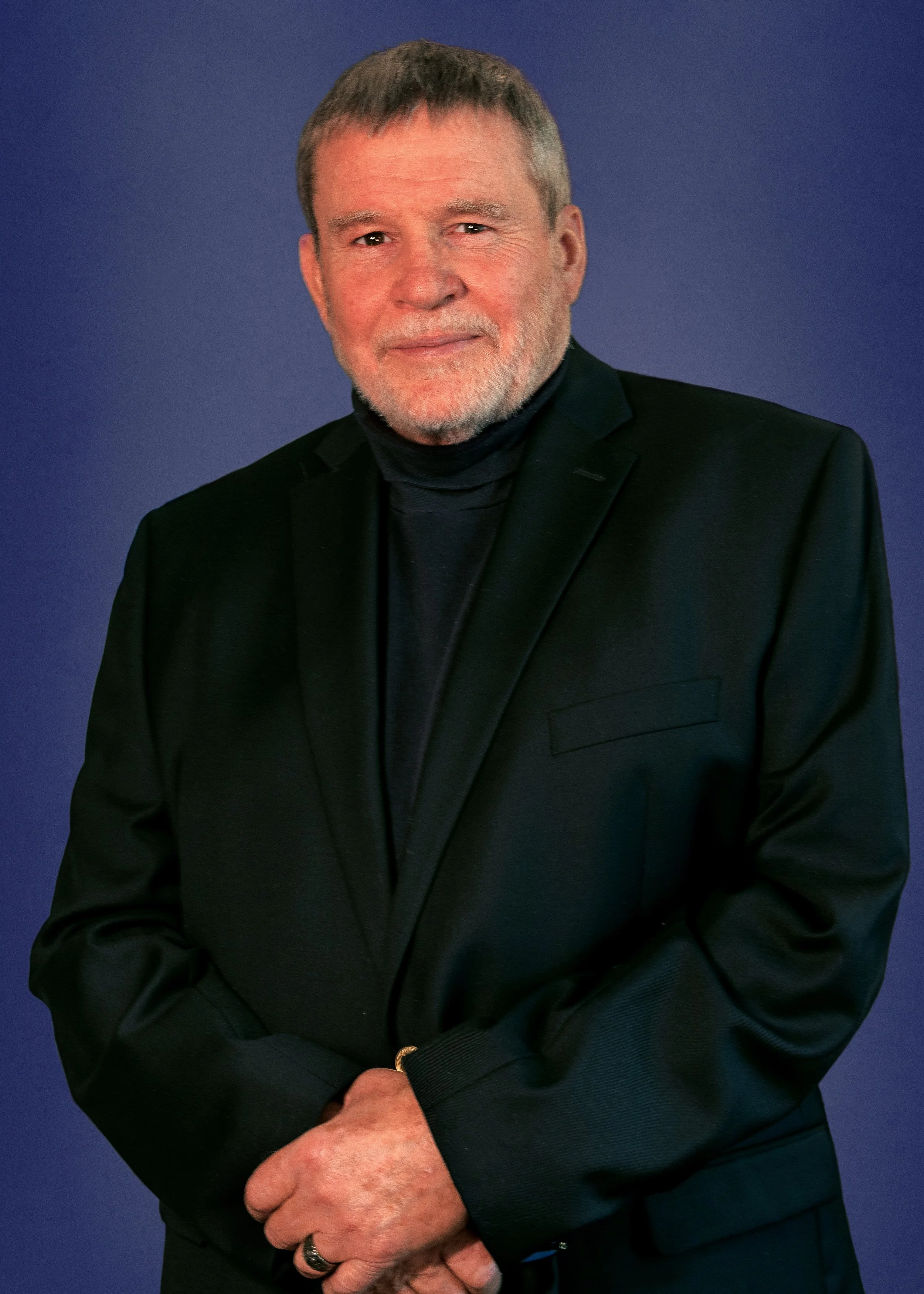 A man with gray hair, beard, and mustache, dressed in a black suit and black turtleneck, standing against a blue background.
