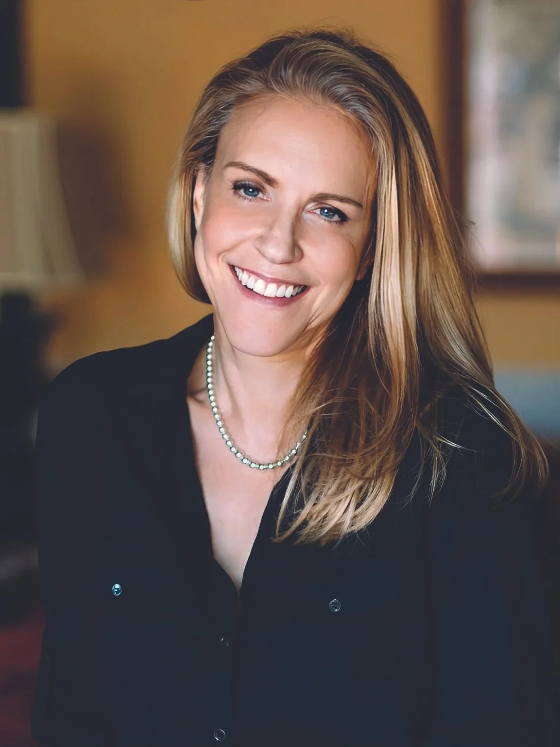 A woman with long blonde hair, wearing a black top and pearl necklace, smiling in a warmly lit room.