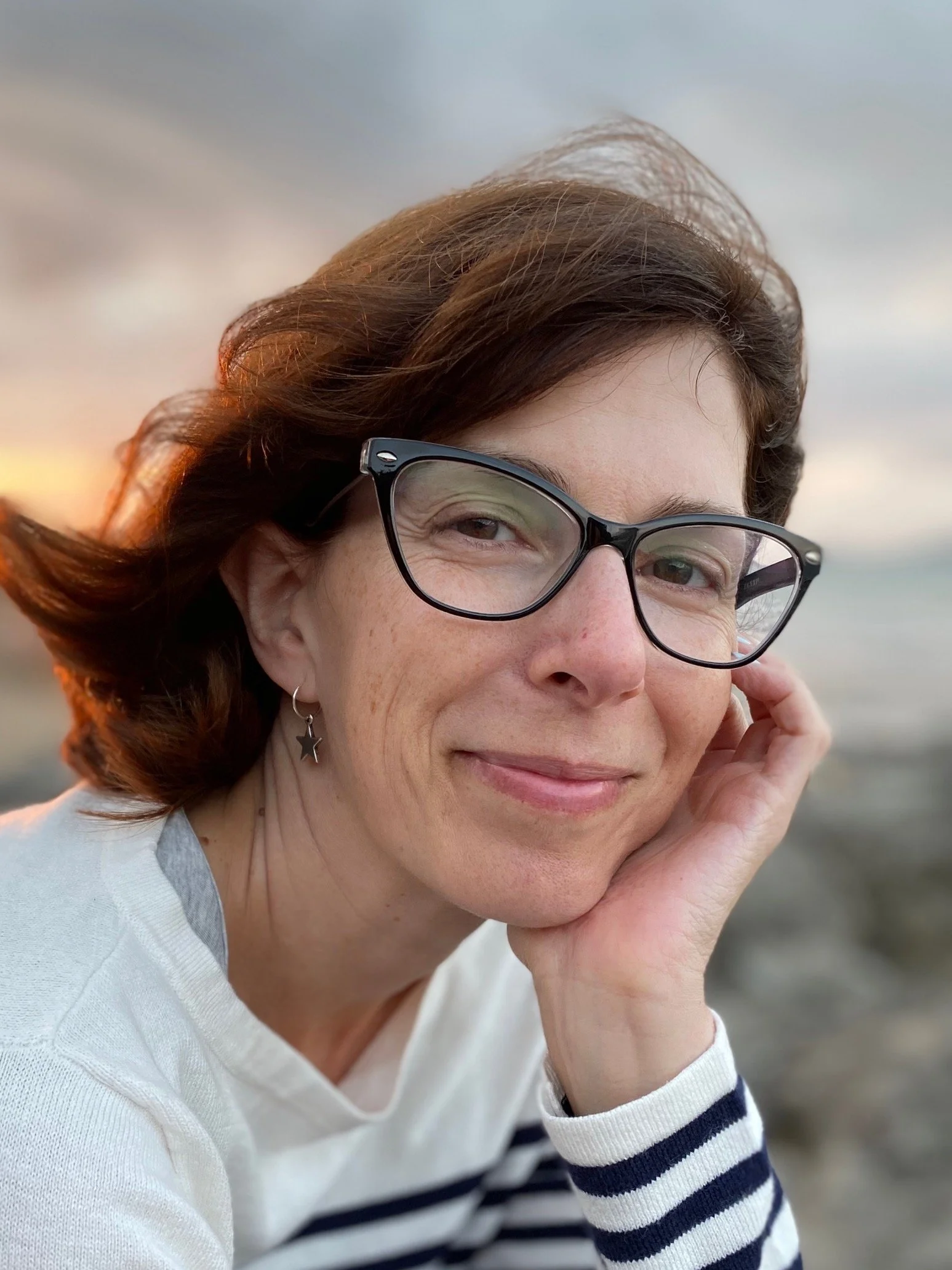 A woman with reddish-brown hair, wearing black glasses and star-shaped earrings, smiling softly at the camera with a hand resting on her cheek, outdoors near a body of water during sunset.