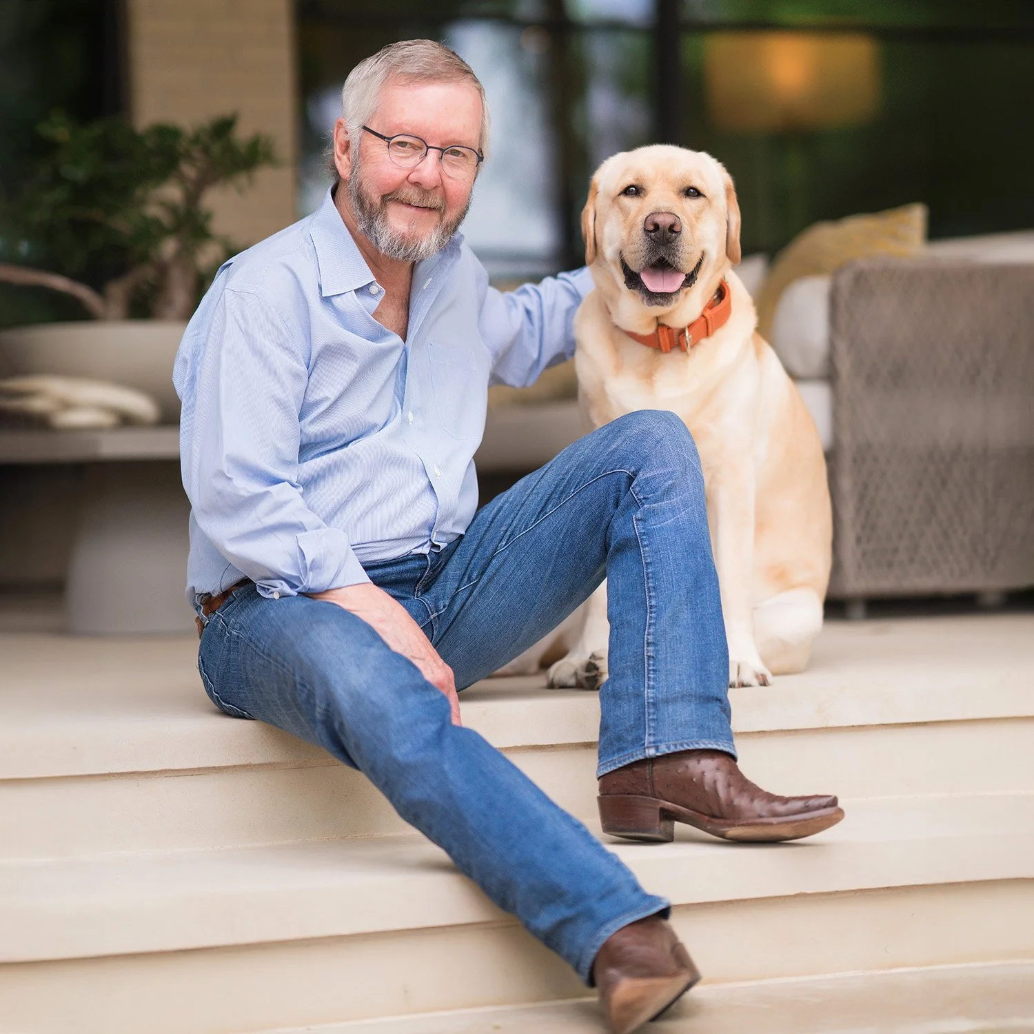A man with gray hair and glasses sitting on steps with a yellow Labrador retriever dog. The man is smiling and wearing a light blue button-down shirt, blue jeans, and brown cowboy boots, with his arm around the dog. The dog is sitting beside him, facing forward with a happy expression, wearing an orange collar.