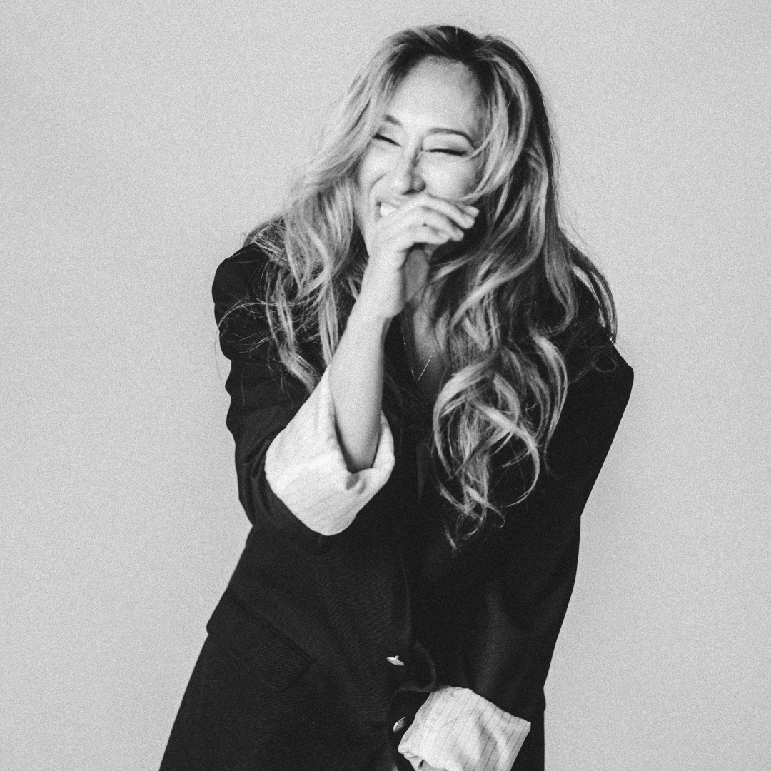 Black and white photo of a woman laughing with her eyes closed, covering her mouth with her hand, with long wavy hair, wearing a dark blazer and striped shirt.