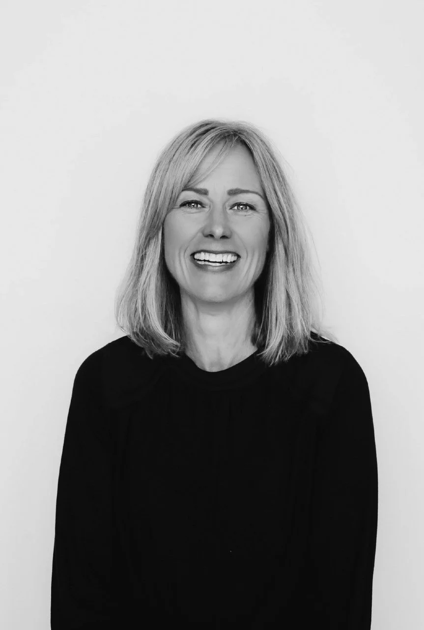 Black and white portrait of a smiling woman with shoulder-length blonde hair, wearing a dark top, against a plain light background.