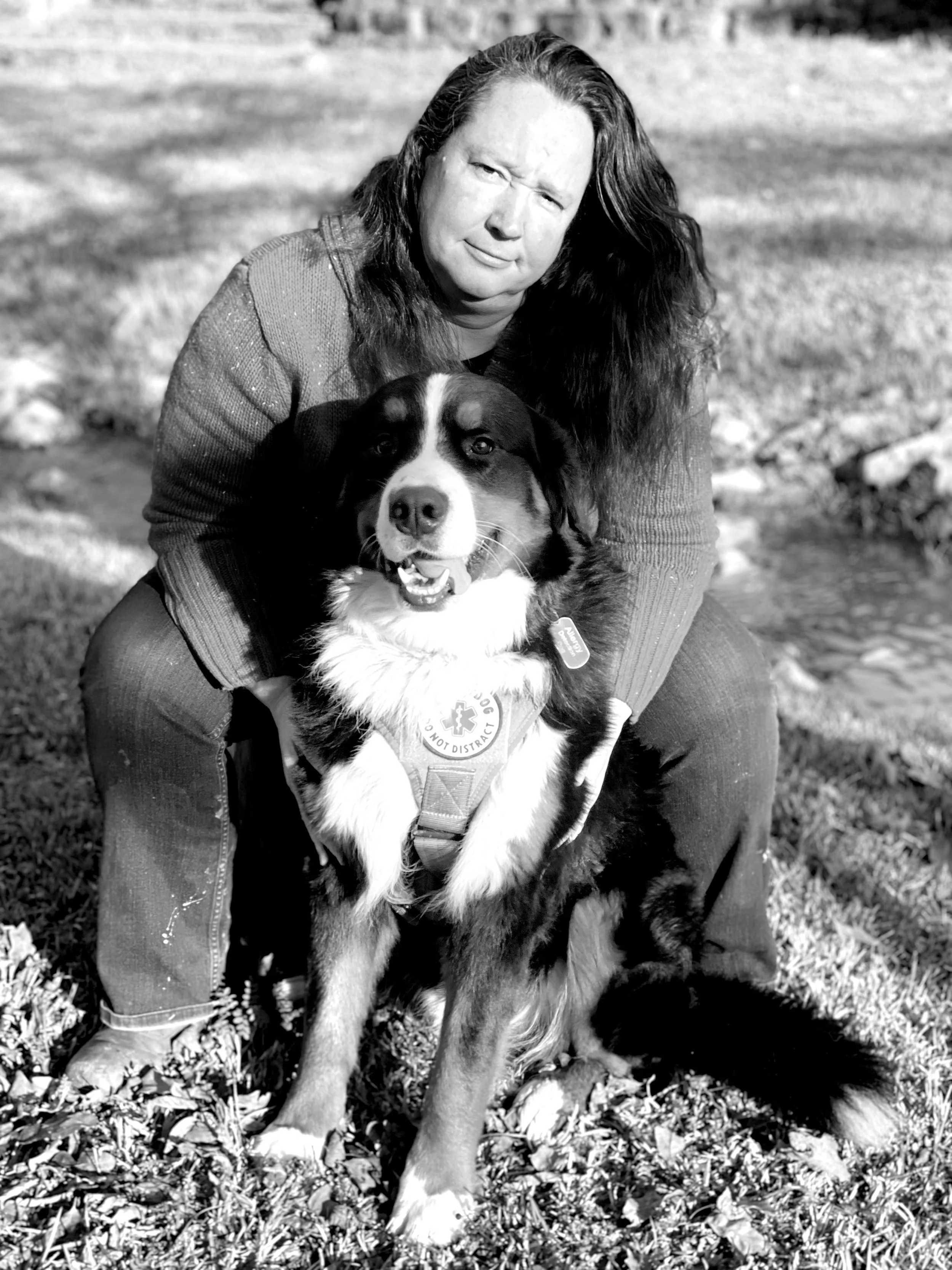 A woman crouching outdoors with a large, happy dog wearing a service vest.