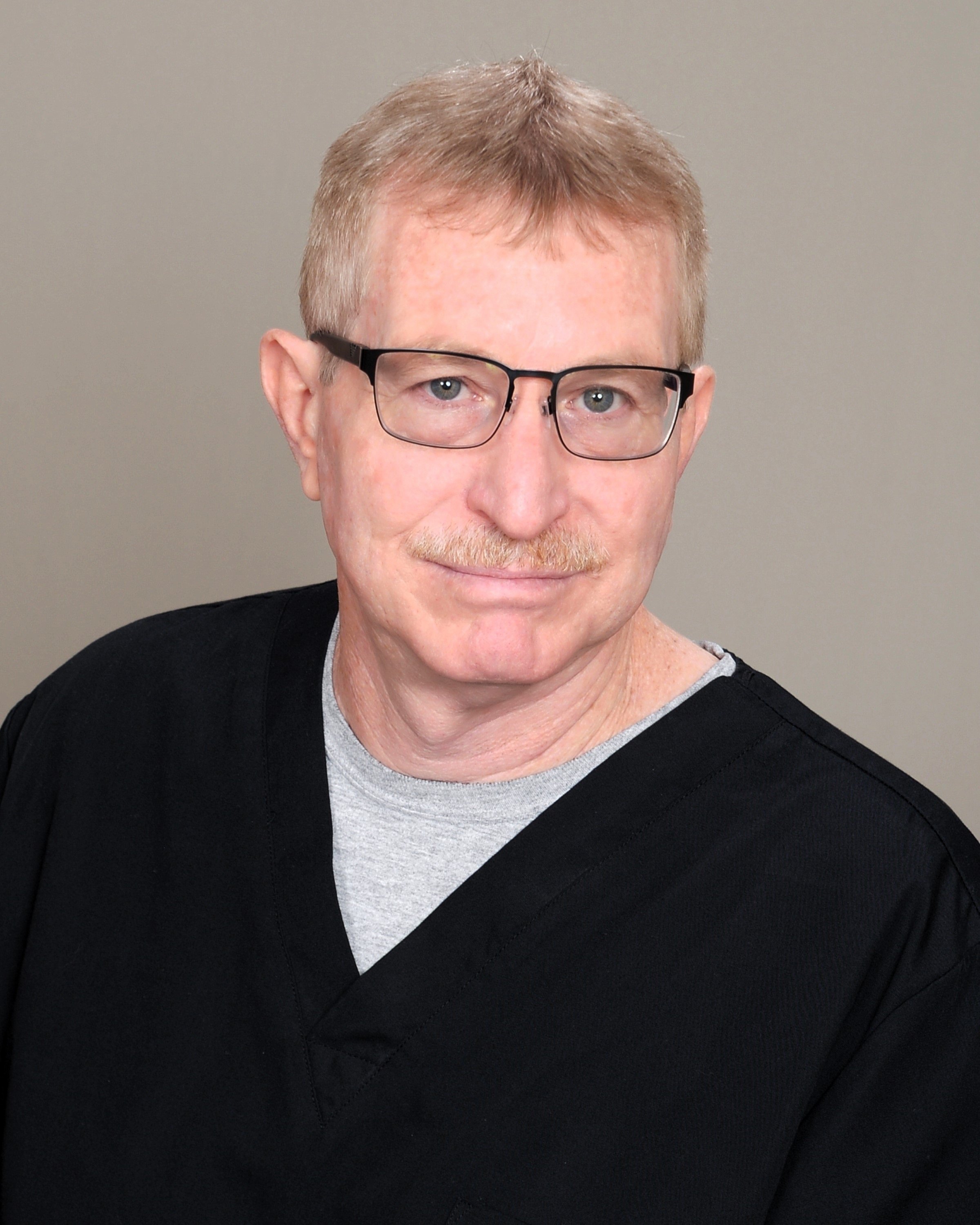 A middle-aged man with light hair, wearing glasses, a gray shirt, and a black sweater, posing against a plain background.