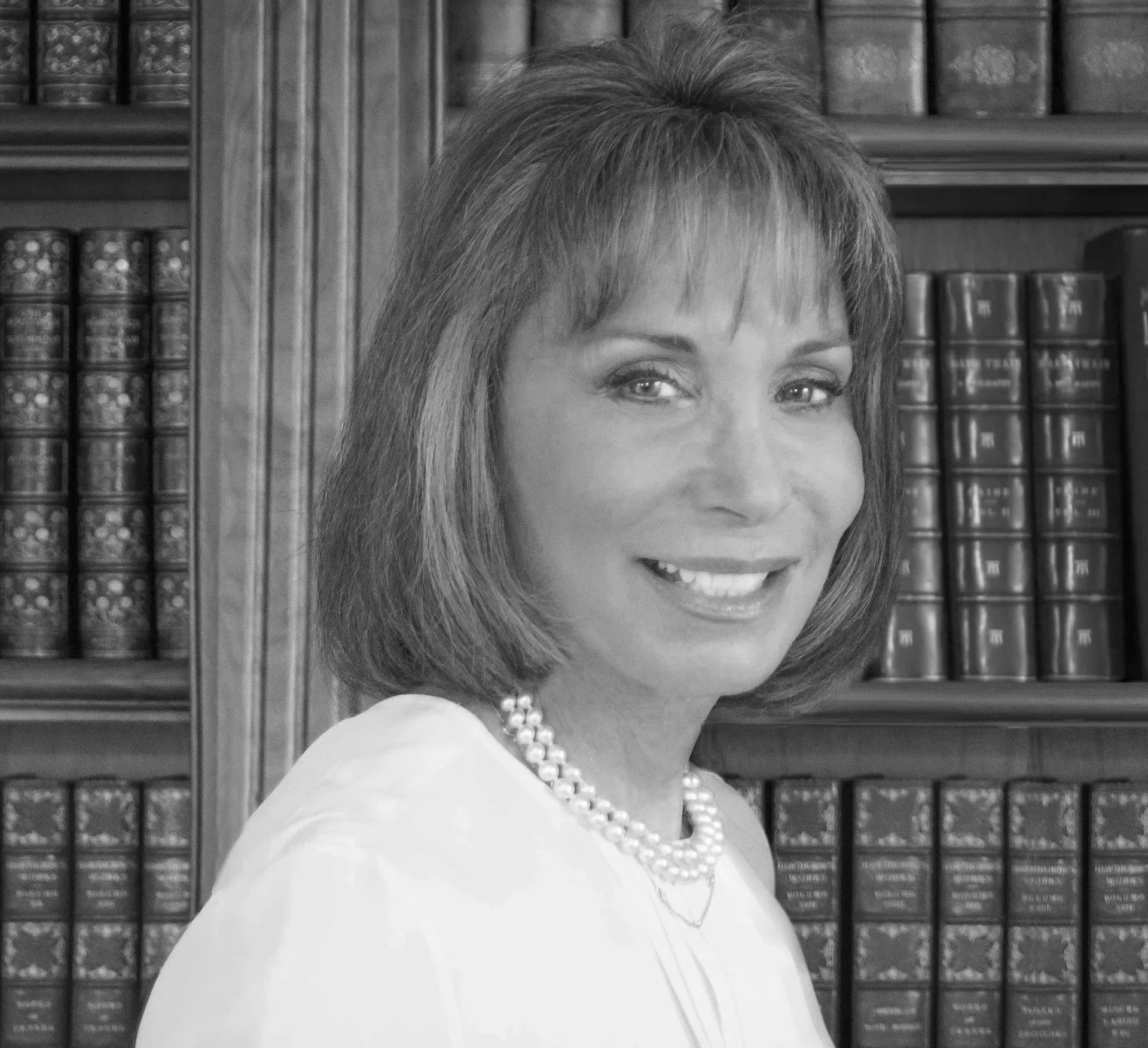 A woman with shoulder-length hair smiling and wearing a pearl necklace, standing in front of a bookshelf filled with leather-bound books.