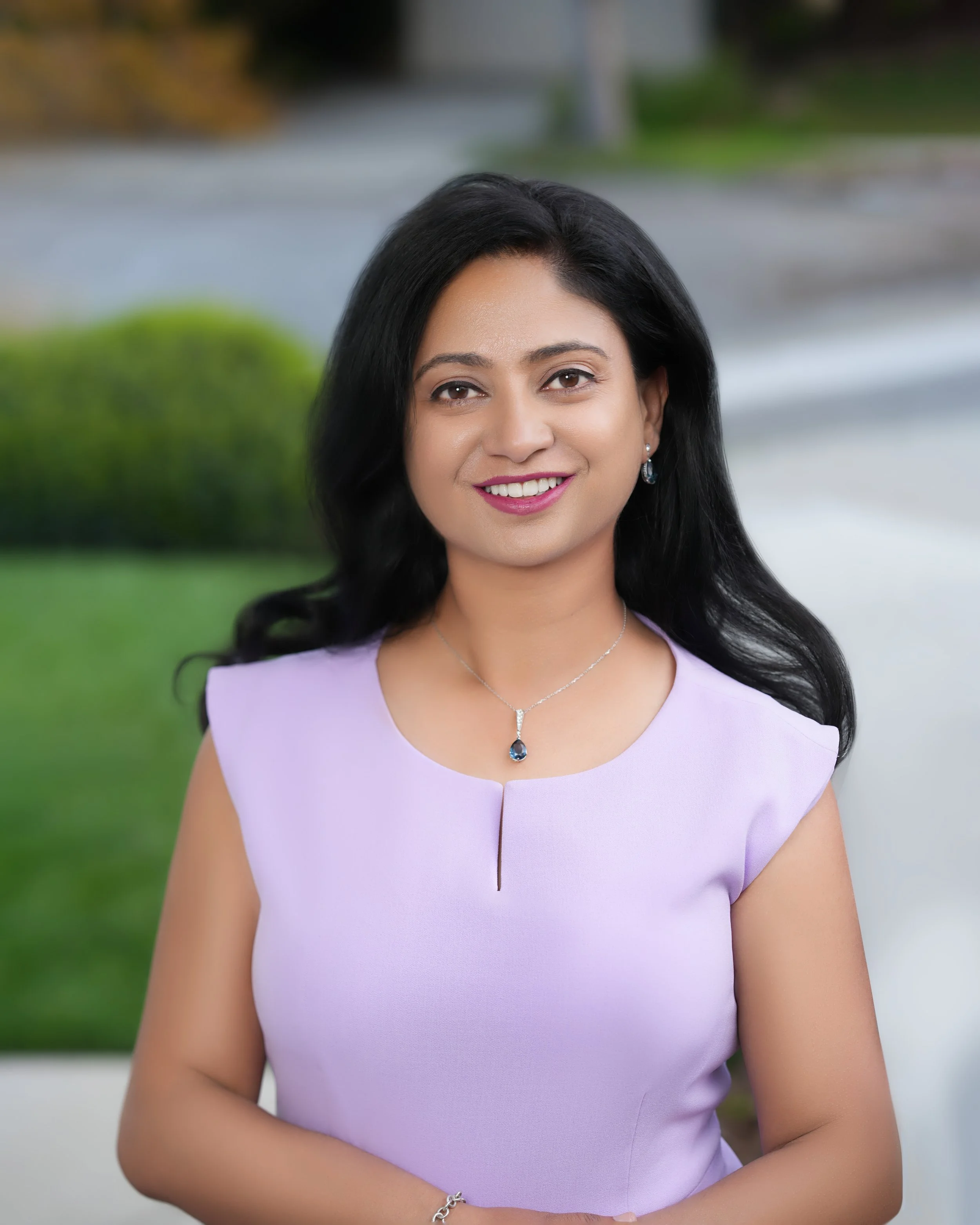 A woman with long black hair wearing a light purple sleeveless dress and jewelry, standing outdoors with greenery and a sidewalk in the background.