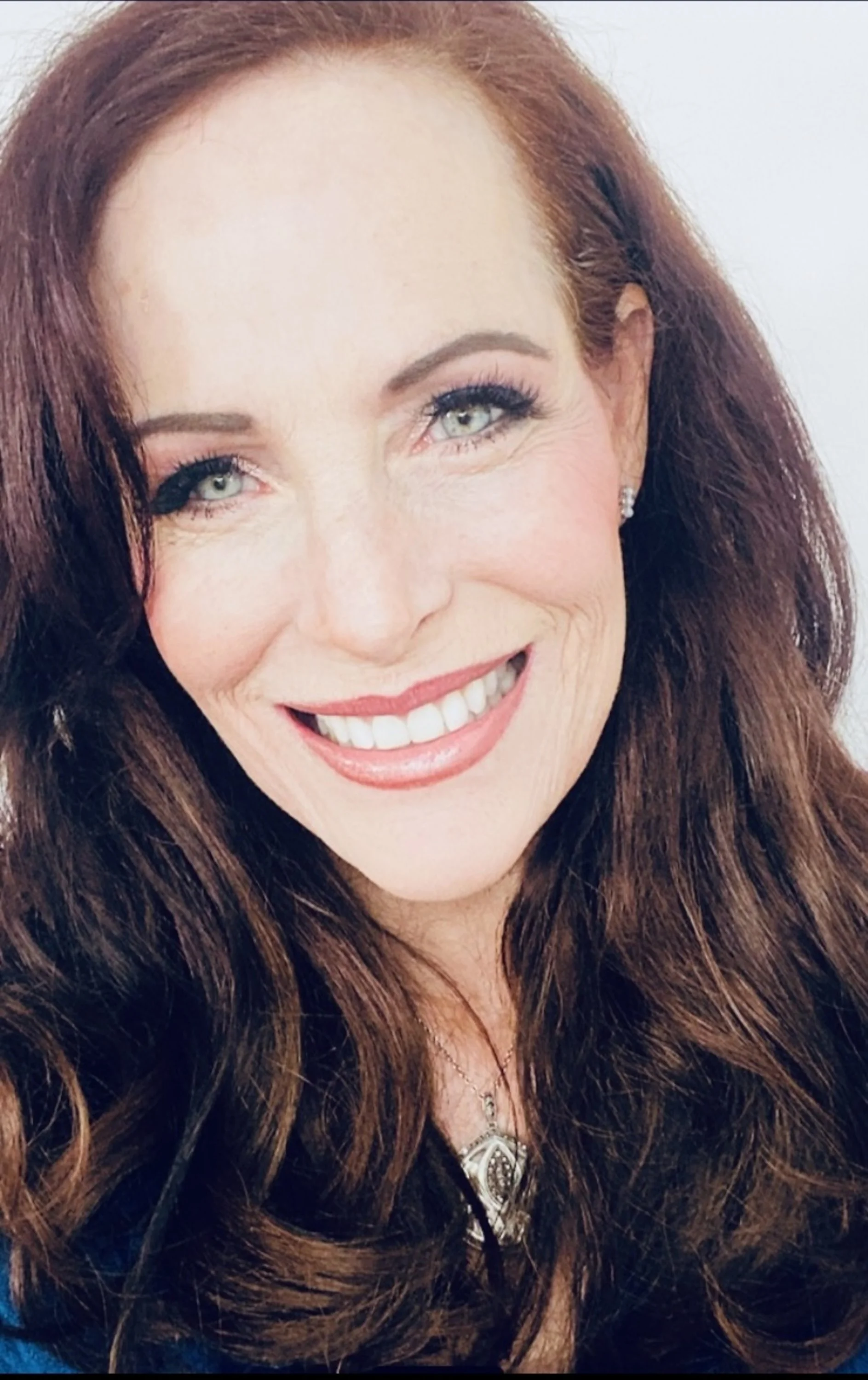 A woman with long, wavy auburn hair and fair skin smiling showing her teeth, wearing earrings and a silver necklace.