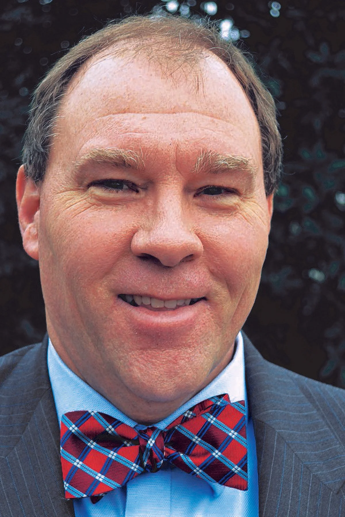 A man with light skin, wearing a blue dress shirt, a red plaid bow tie, and a dark suit, smiling outdoors with dark foliage in the background.