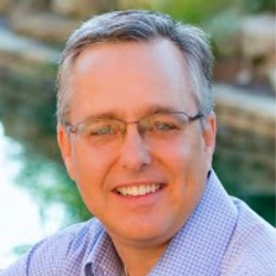 A middle-aged man with glasses and gray hair smiling outdoors with water and rocks in the background.