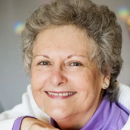 Close-up of an elderly woman smiling, with short curly gray hair, wearing a white and purple shirt, and small earrings.