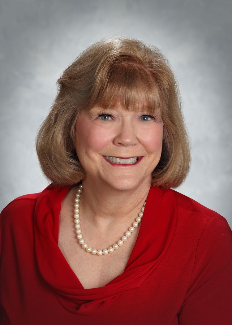 A smiling elderly woman with shoulder-length light brown hair, wearing a red blouse and a string of white pearls, against a gray background.