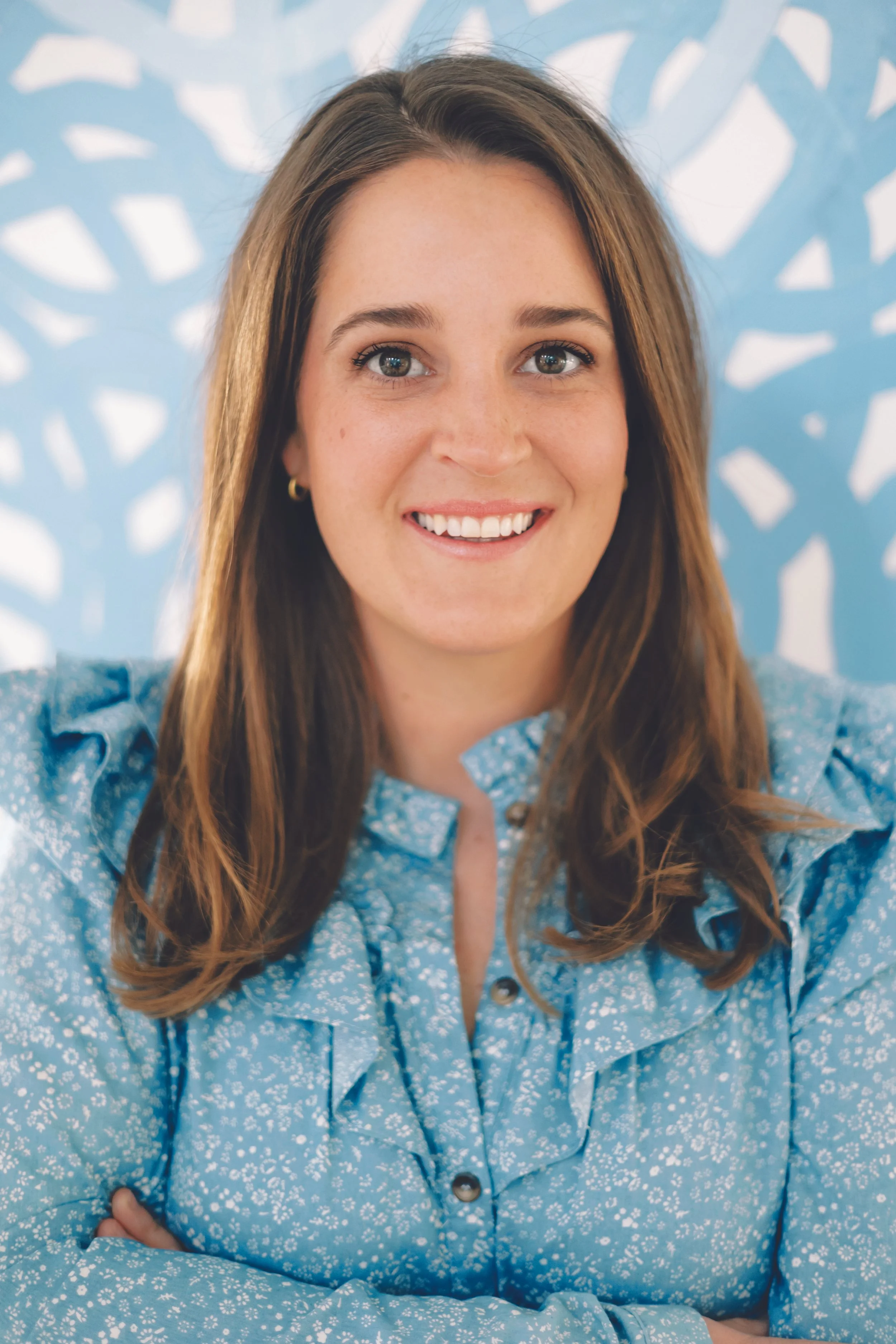 A woman smiling in front of a blue patterned background, wearing a blue floral blouse.