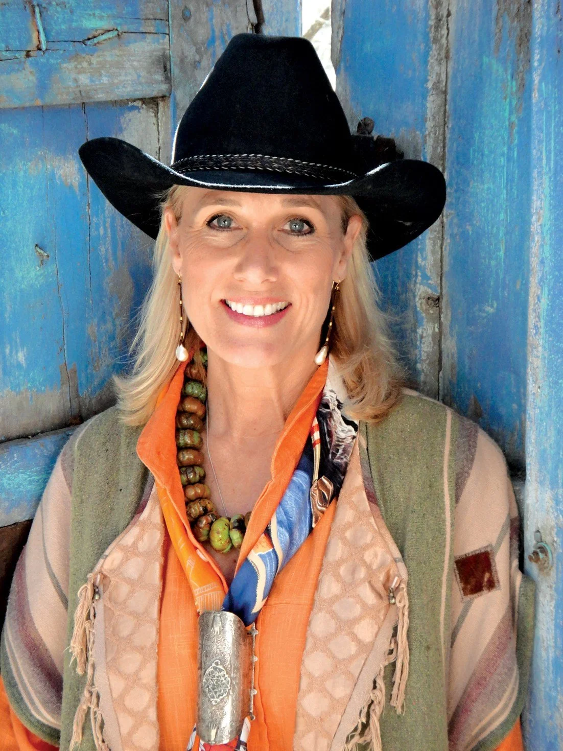 A woman smiling, wearing a black cowboy hat, colorful scarf, and beaded necklace, standing in front of a rustic blue wooden background.