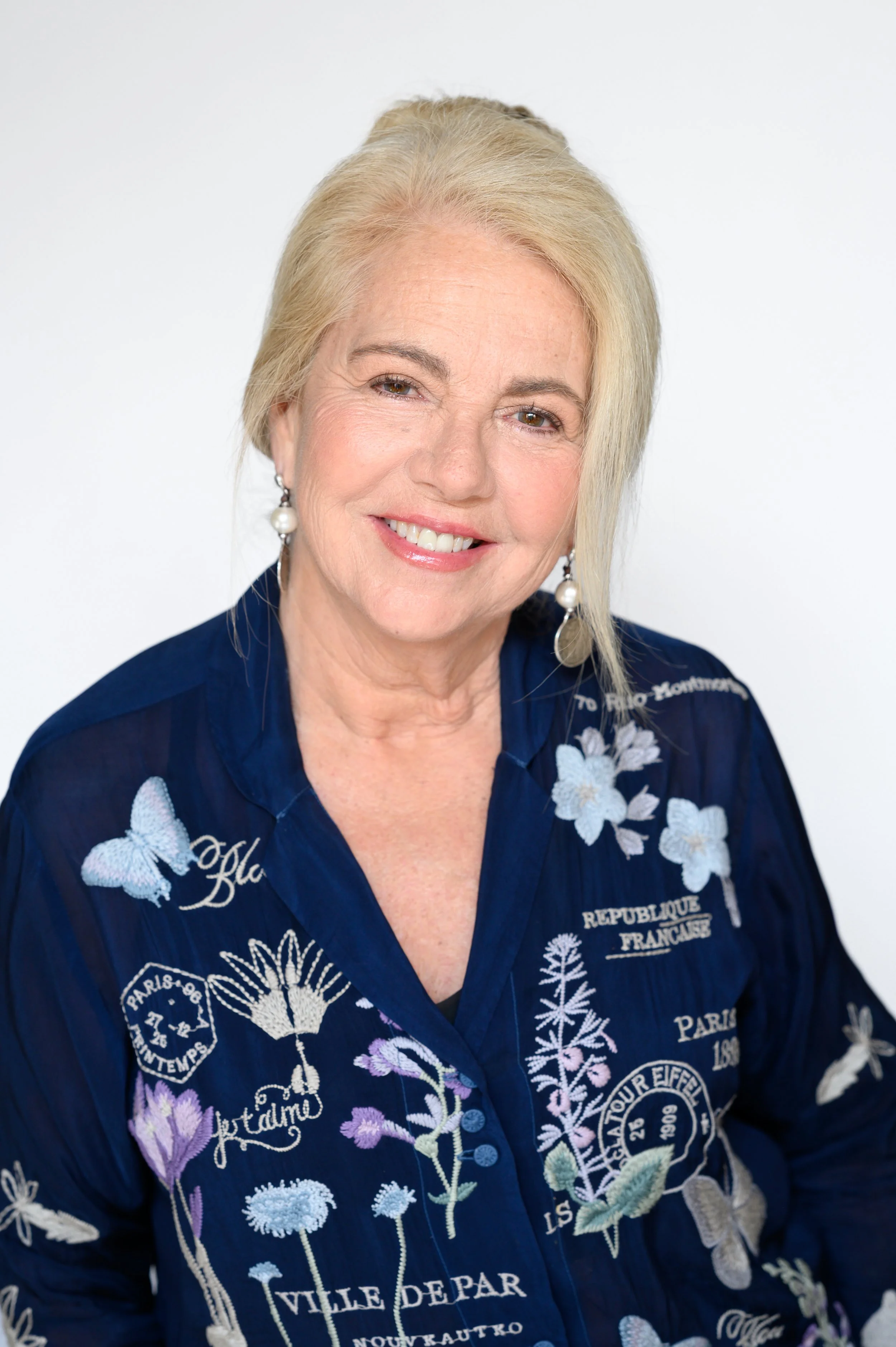 An older woman with blonde hair smiling, wearing a navy blue embroidered shirt with floral and butterfly designs and pearl earrings.