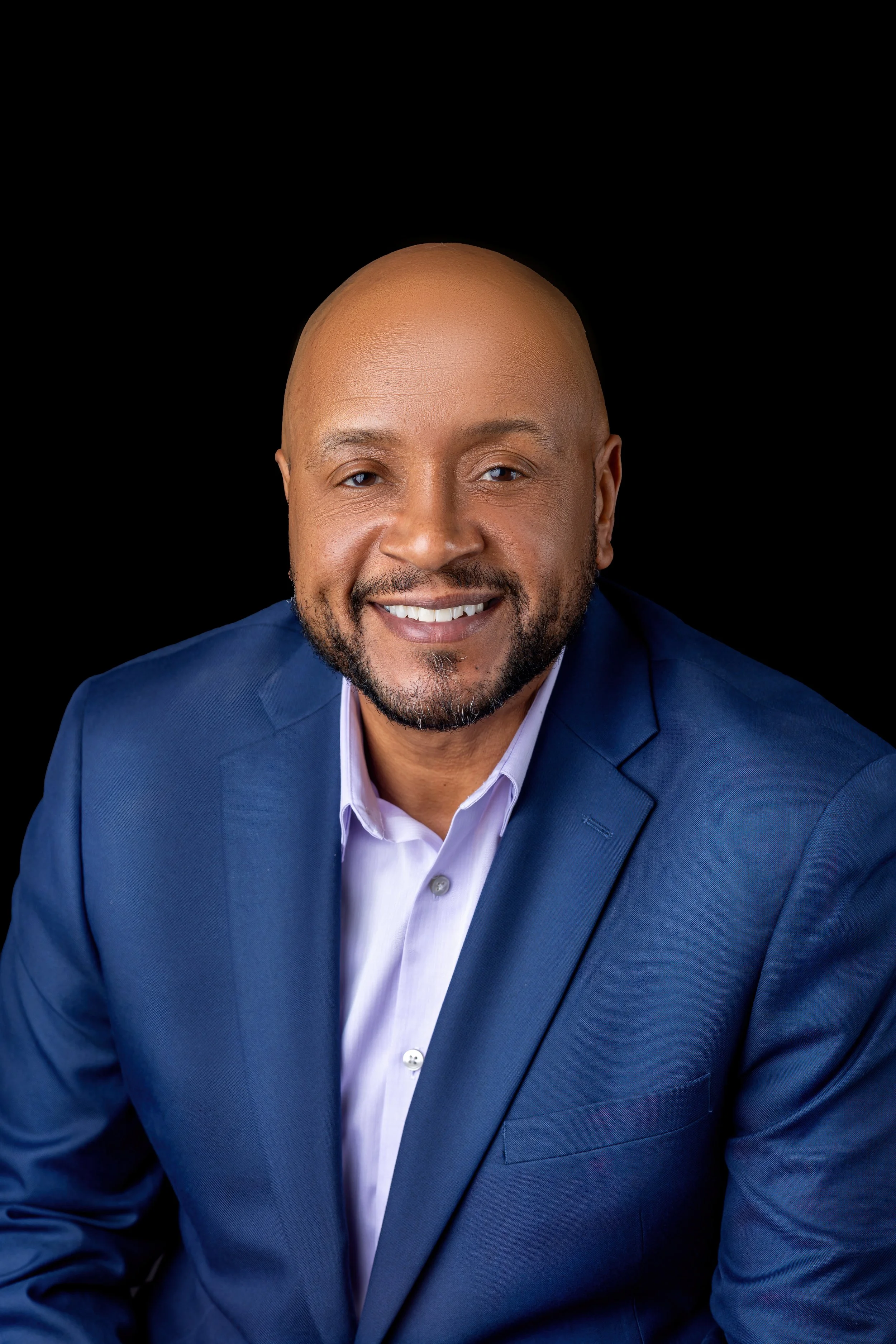 Portrait of a smiling African American man with a beard, in a blue suit and white shirt, against a black background.