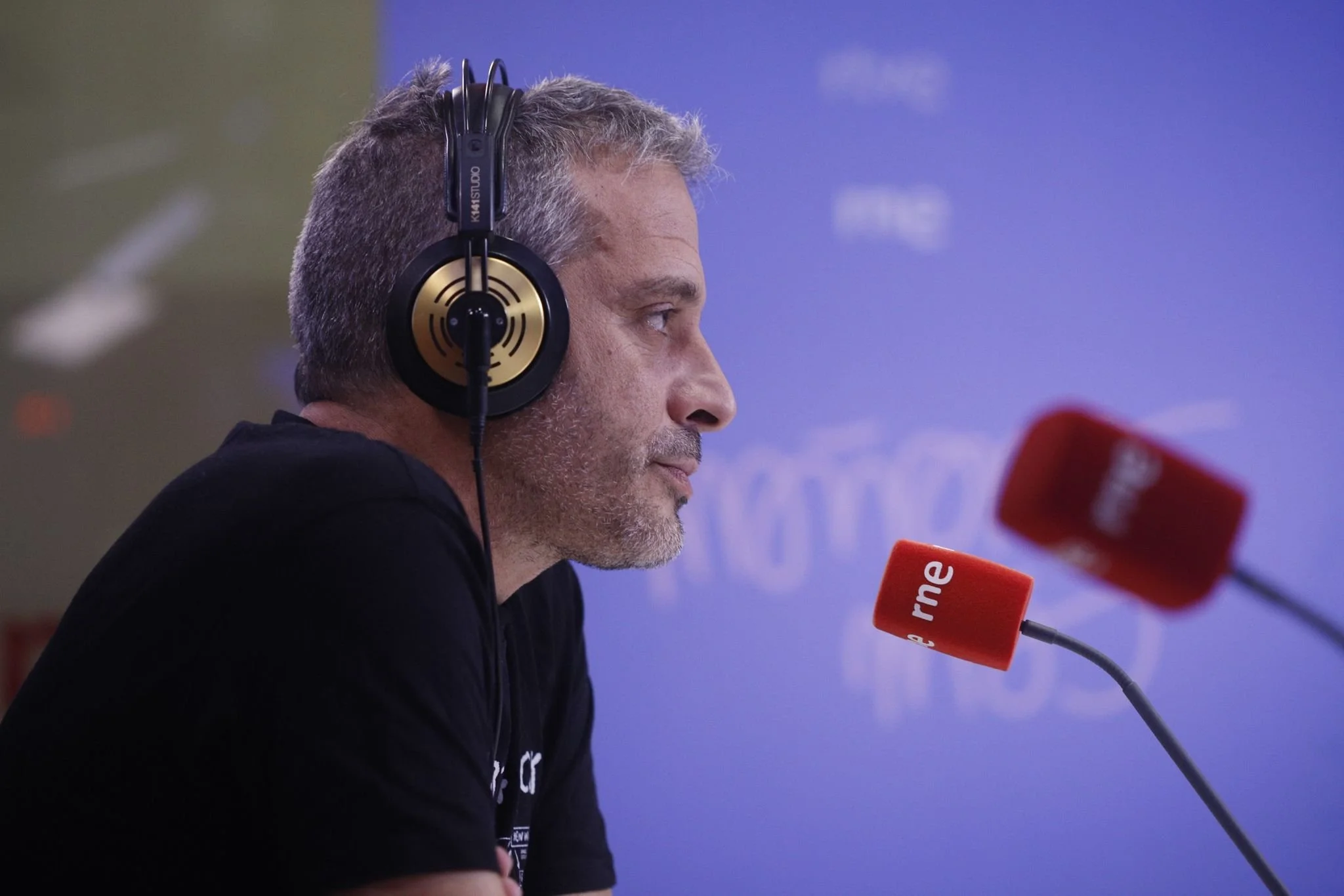 A man with gray hair and beard wearing headphones, sitting in front of a microphone labeled 'rne', inside a radio station or studio.