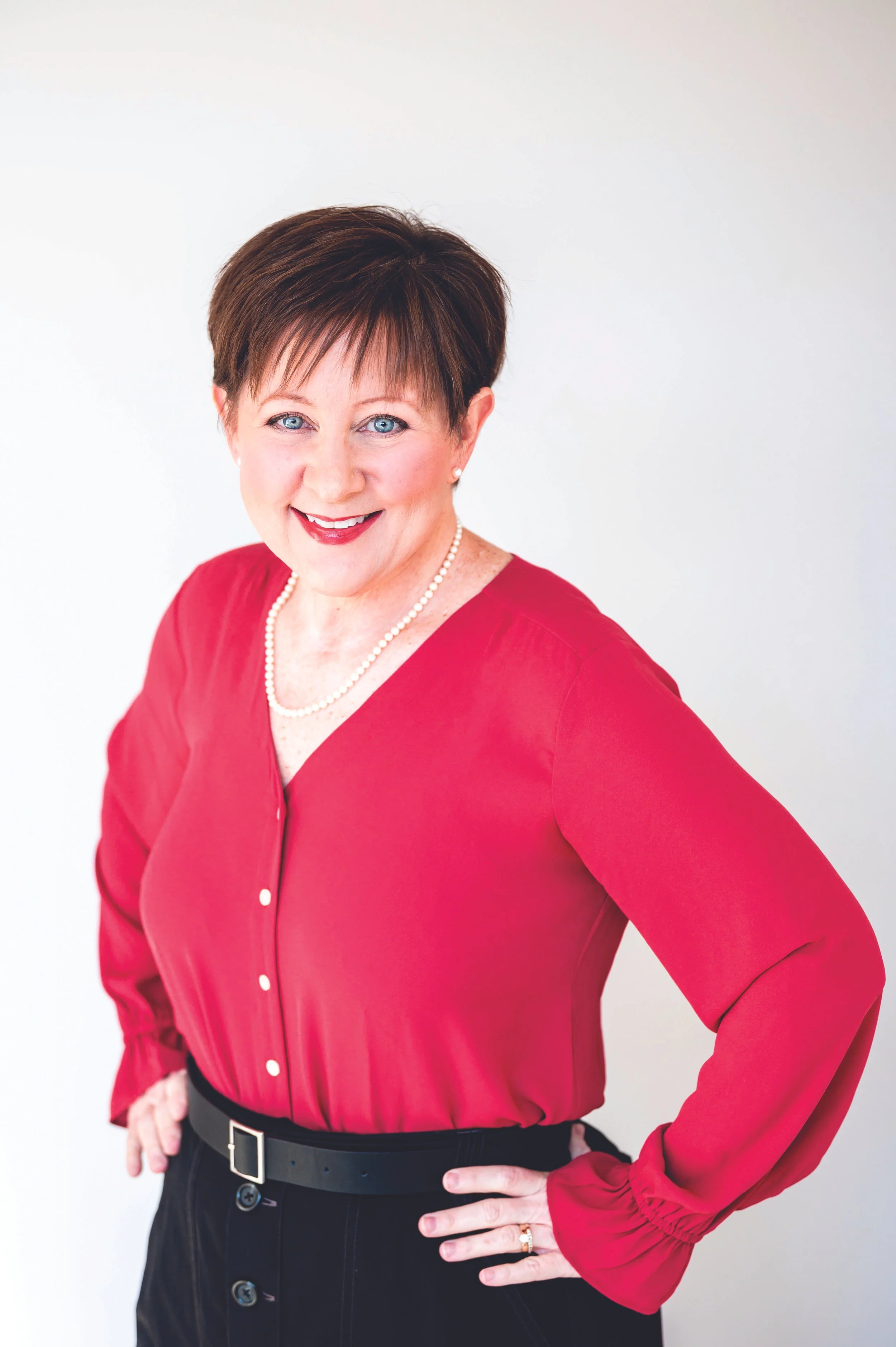 A smiling middle-aged woman with short brown hair and blue eyes, wearing a red blouse, black pants, a pearl necklace, and a ring, standing against a plain white background.