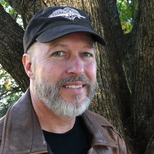 Man with a gray beard, wearing a black cap with white text and an emblem, a brown leather jacket, and a black shirt, standing outdoors in front of a tree with green foliage.