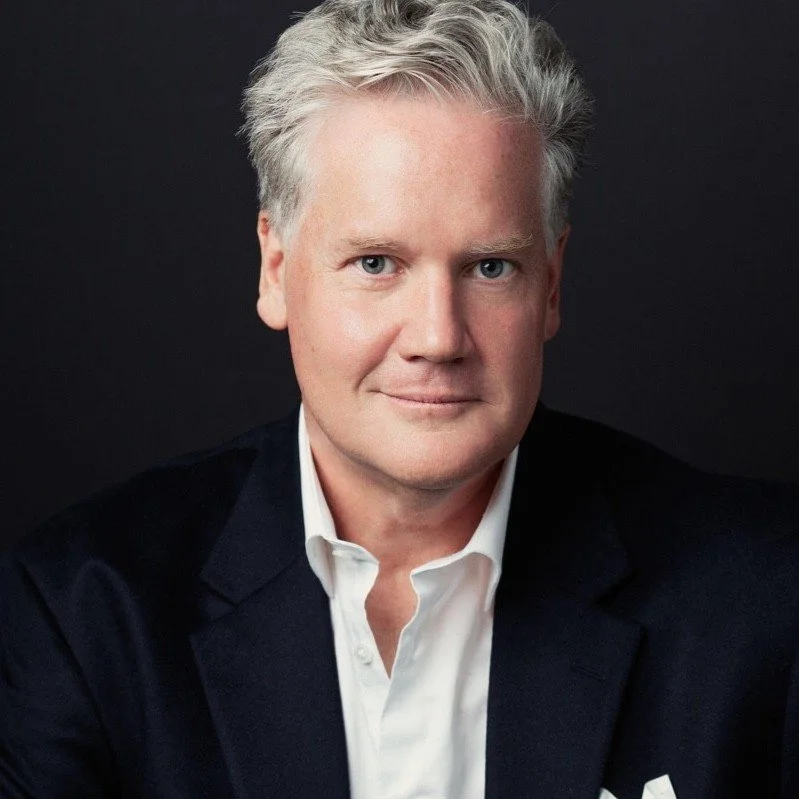 A professional headshot of a middle-aged man with light skin, gray hair, wearing a dark blazer and white shirt, against a black background.