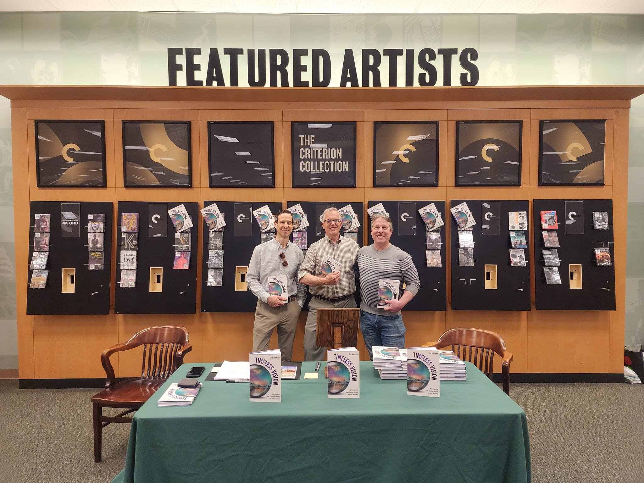 Three men standing behind a table at a book signing event, holding copies of a book titled 'Timeless Vision,' with a backdrop of vending machines and a 'Featured Artists' sign.
