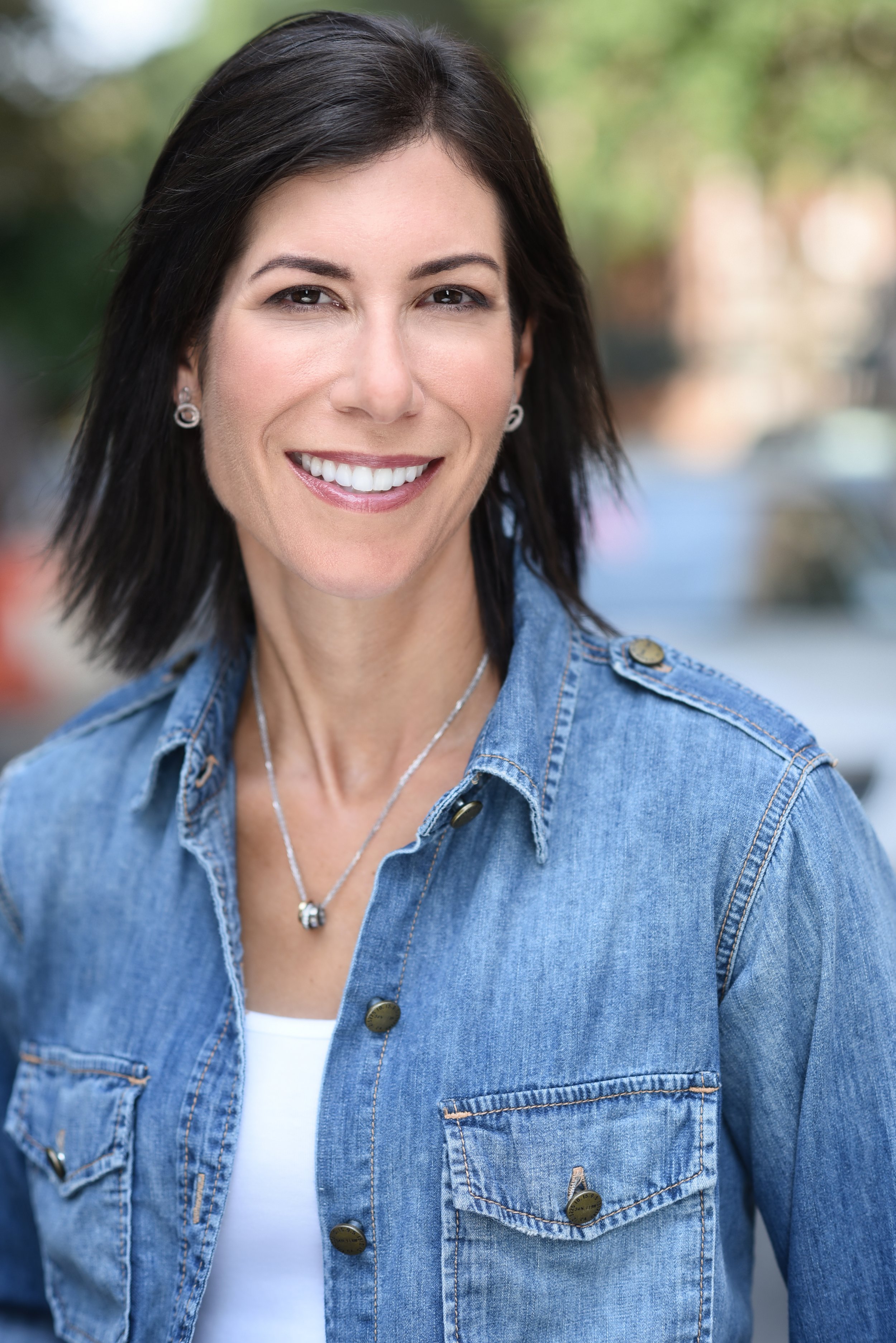 A woman smiling outdoors, wearing a denim jacket, a white top, and jewelry, with a blurred background.