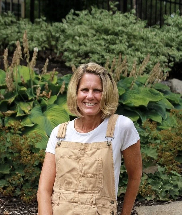 A woman with shoulder-length blonde hair smiling outdoors, wearing a white t-shirt and tan overalls, standing in front of lush green plants.