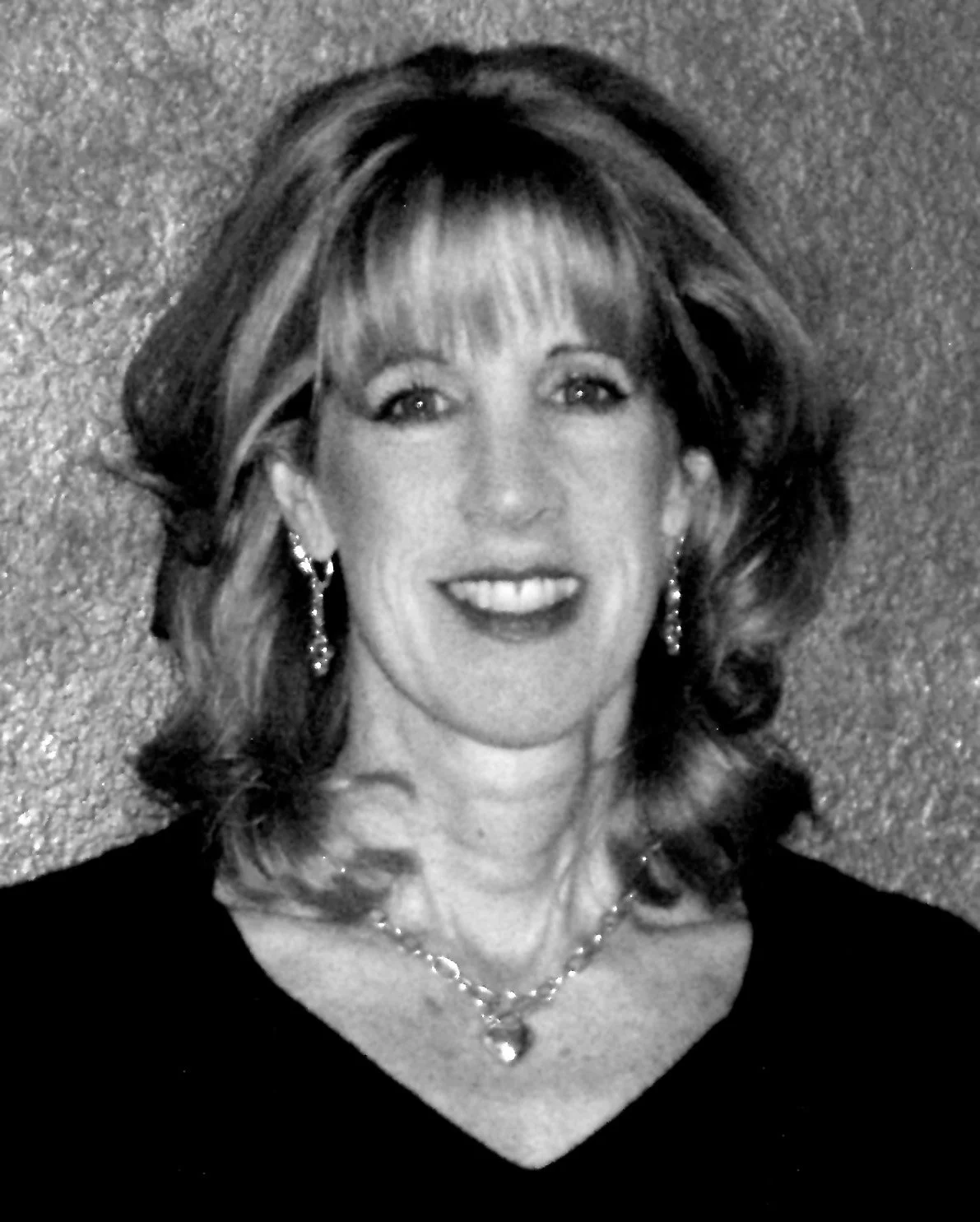 Black and white photograph of a smiling middle-aged woman with shoulder-length hair, wearing earrings, a necklace, and a top with a V-neckline, standing against a textured wall.