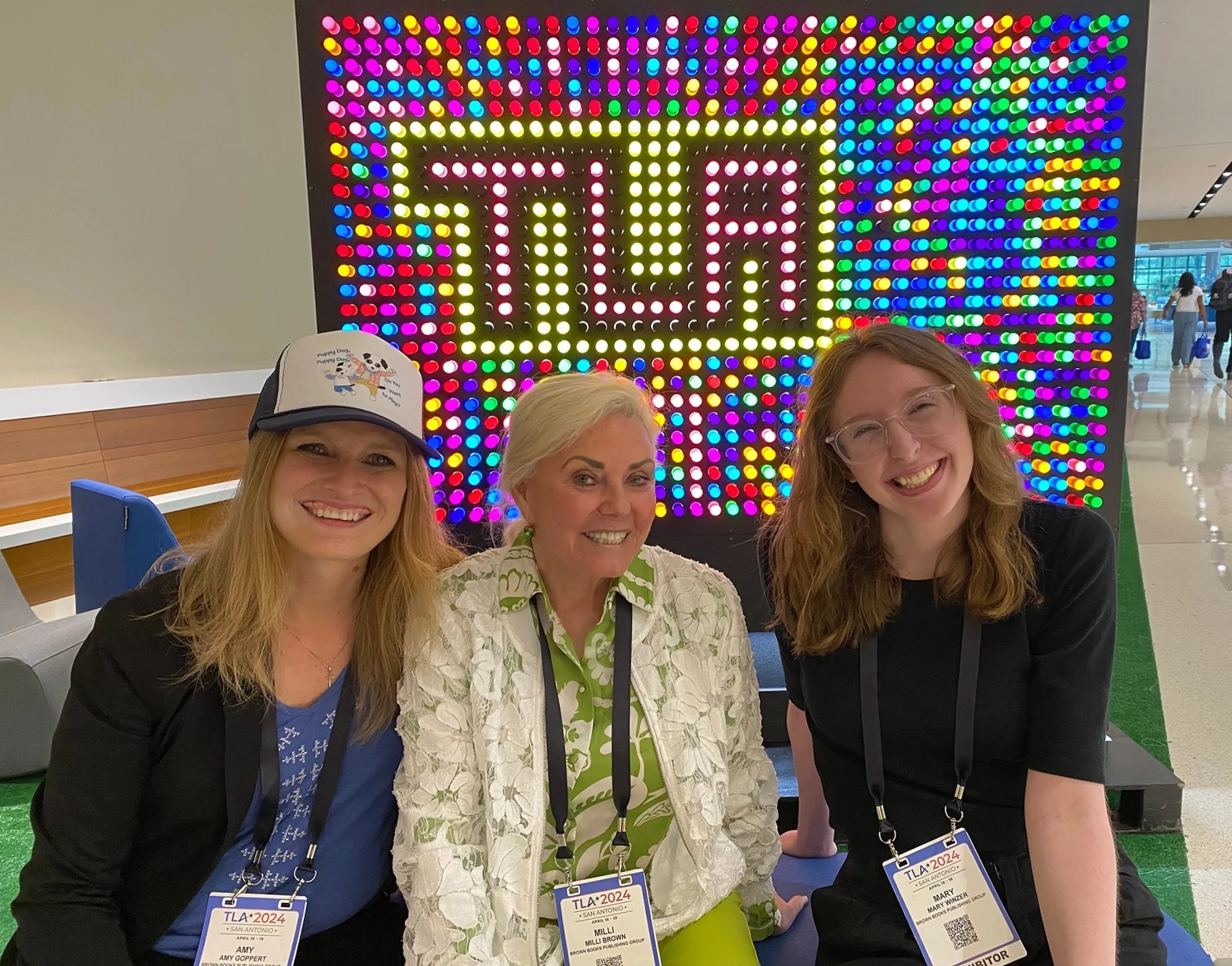 Three women smiling in front of a colorful LED sign that reads "TLA" at a conference. All are wearing name badges and standing indoors.