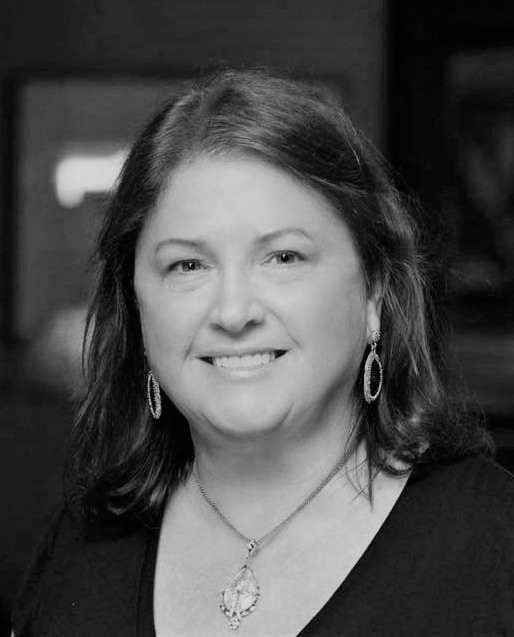 Black and white portrait of a smiling woman with shoulder-length hair, wearing earrings and a necklace, indoors.