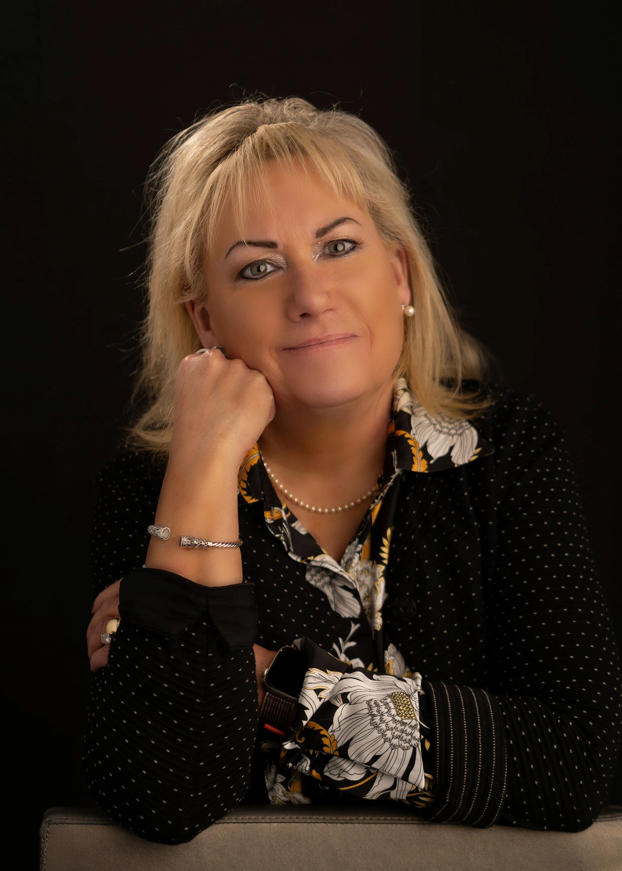 A middle-aged woman with blonde hair, wearing a black and floral patterned blouse, a pearl necklace, and pearl earrings, sitting at a table with her chin resting on her hand against a black background.