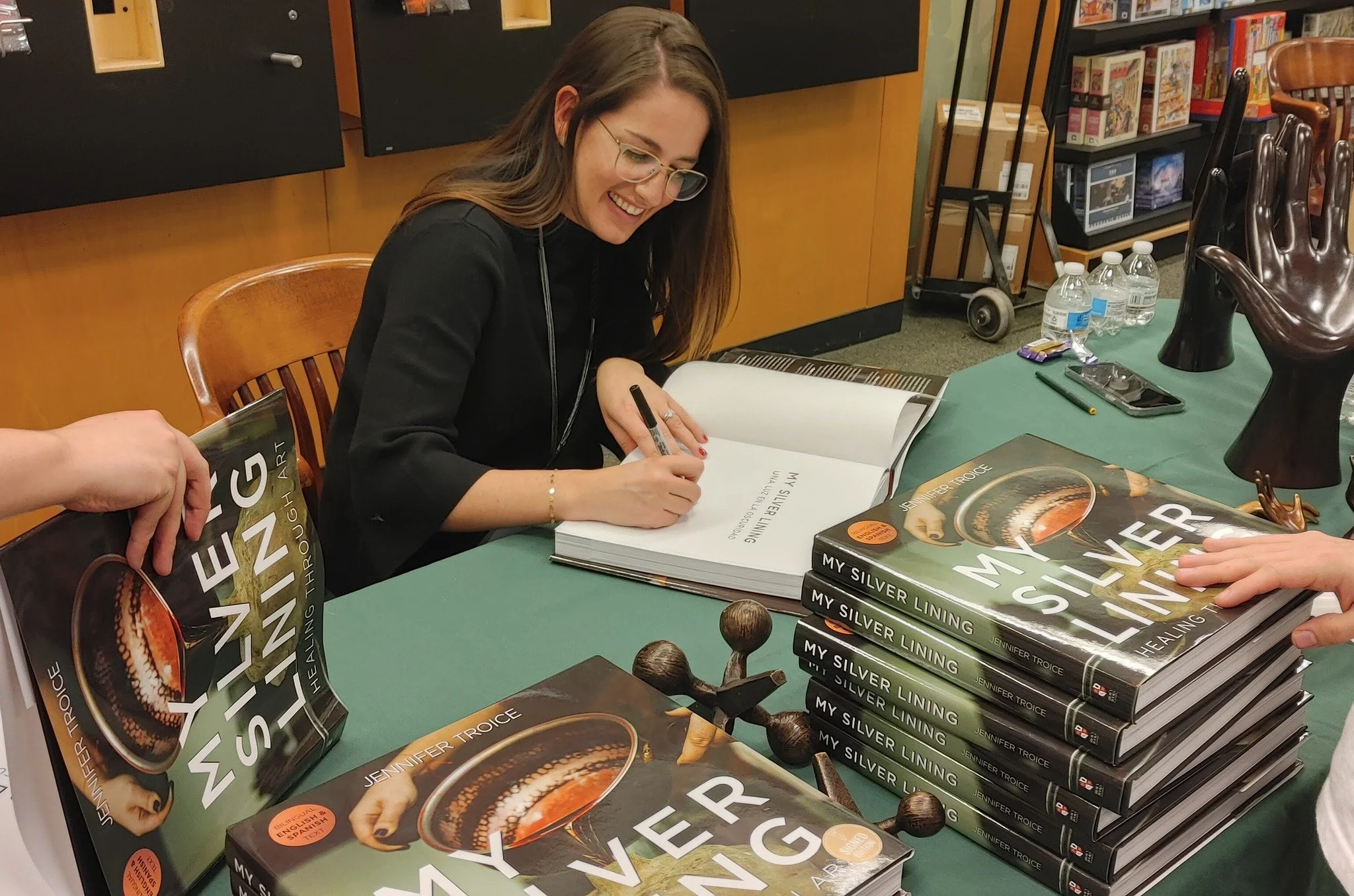 Author Jennifer Troice signing her book titled 'My Silver Lining' at a book event, with stacks of her books on the table and water bottles in the background.