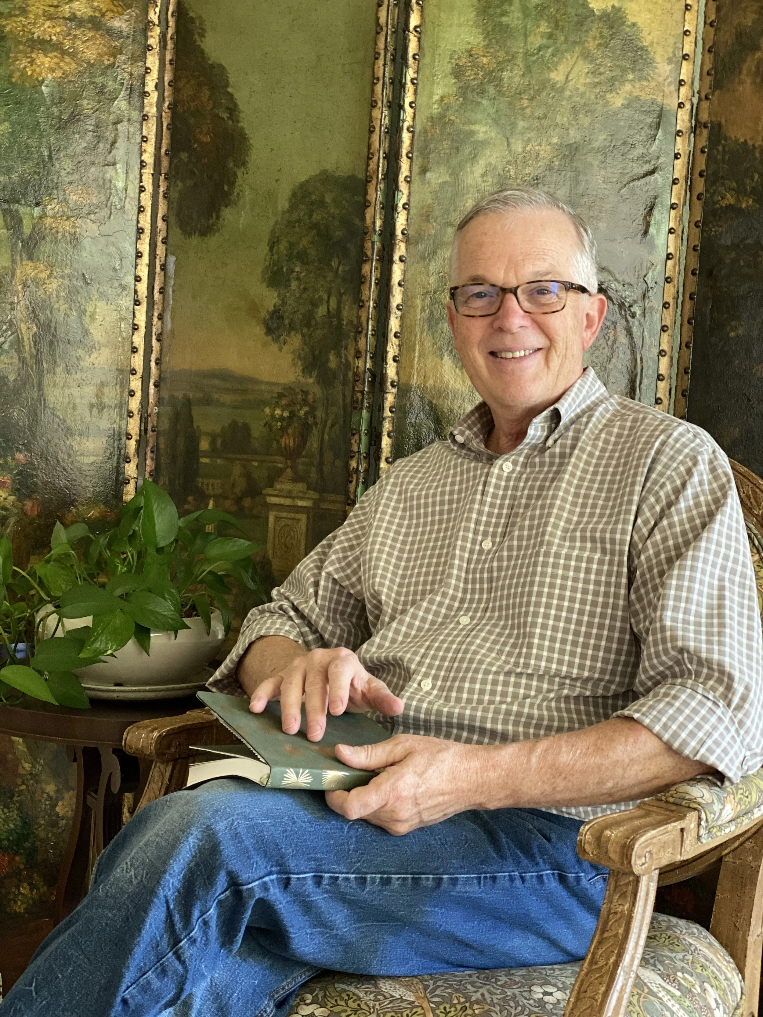 A smiling man with glasses sitting on an ornate chair, holding a book, with a painted landscape backdrop and a potted plant on a side table.