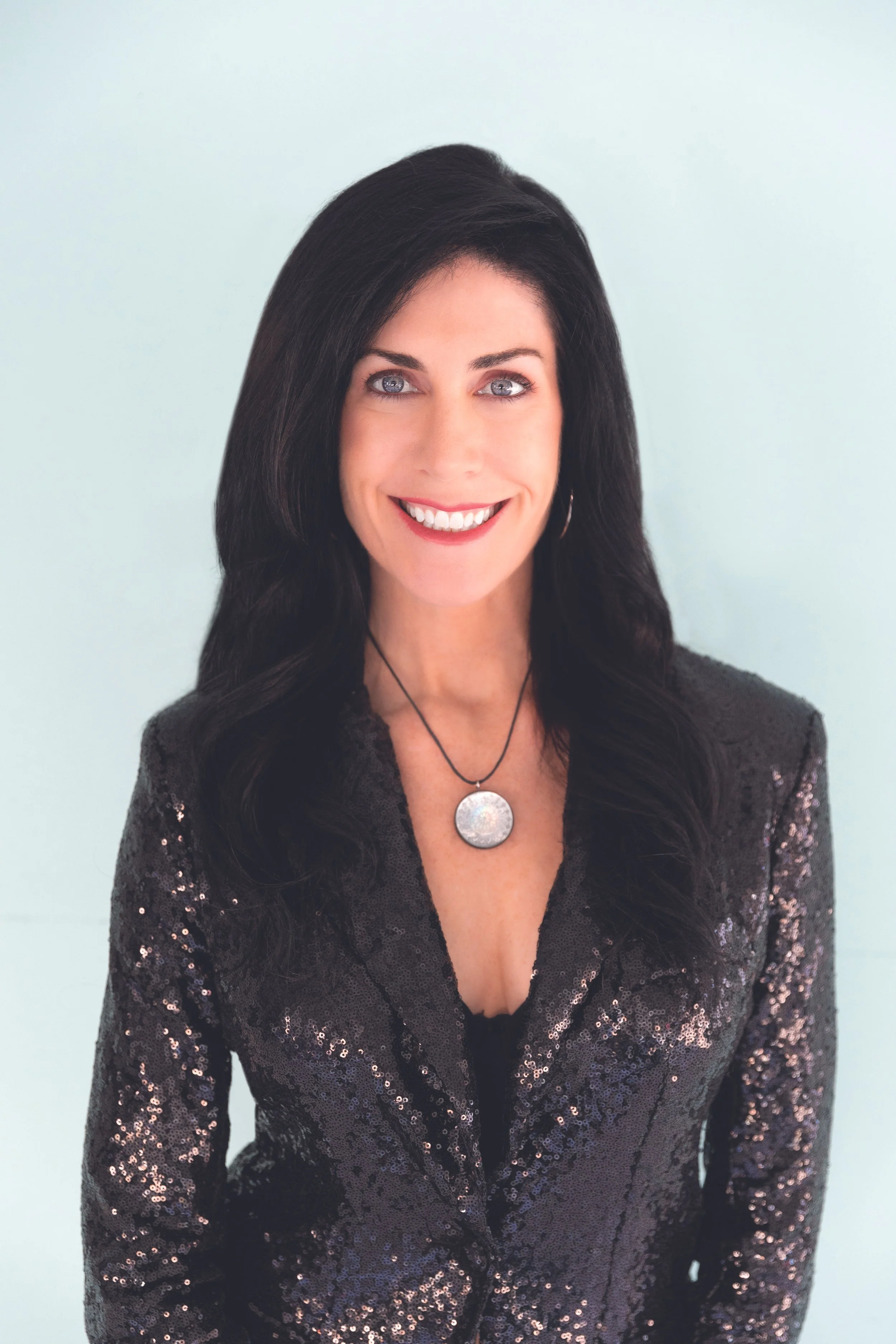A woman with black, wavy hair smiling and wearing a black sequin blazer with a round pendant necklace.