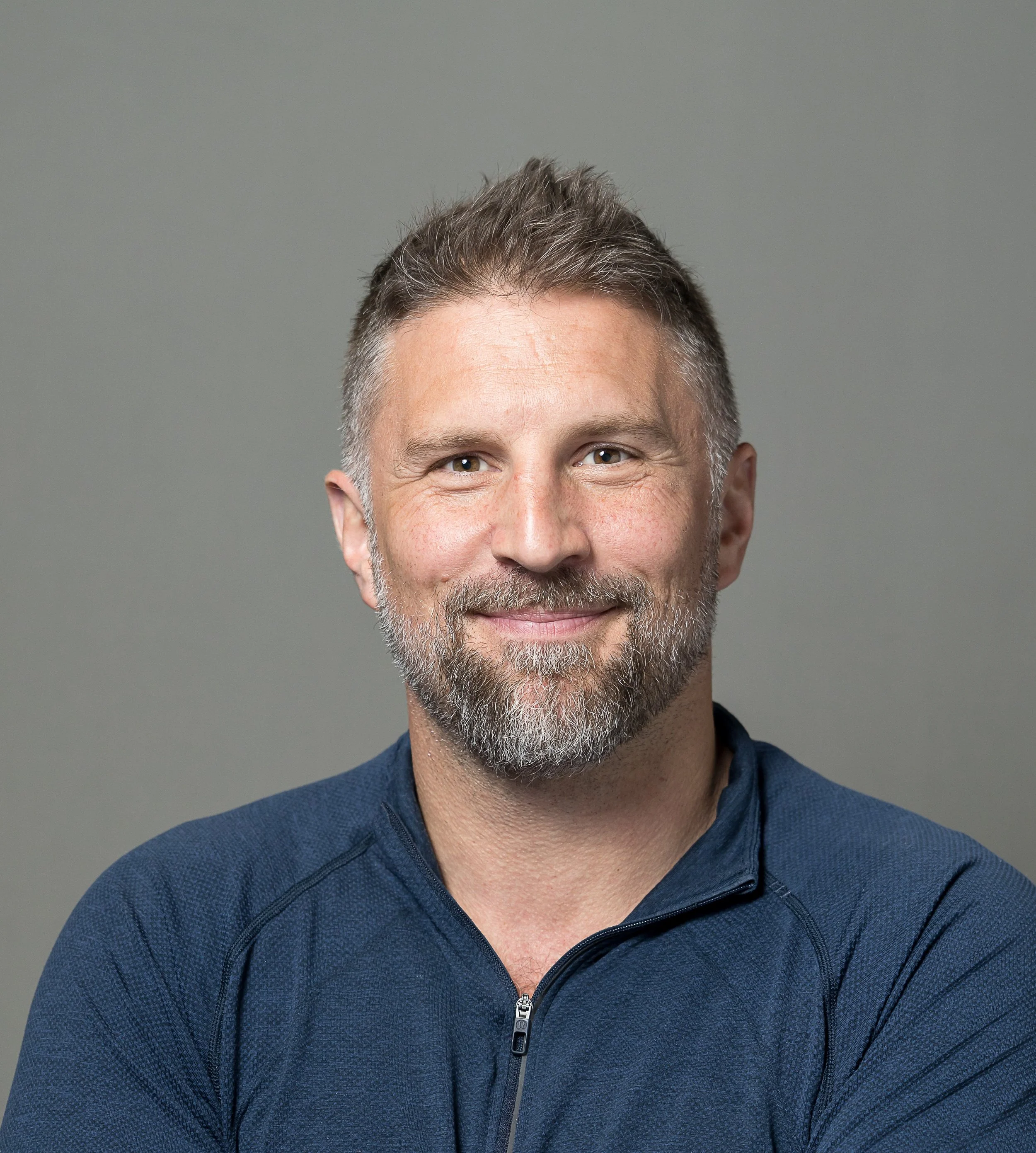 Headshot of a middle-aged man with short gray and brown hair, a beard, and wearing a navy blue zip-up sports jacket, smiling against a plain gray background.