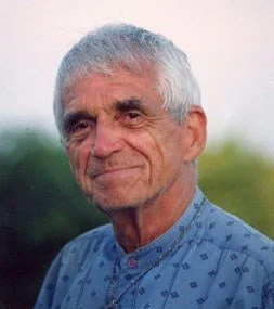 Close-up of an elderly man with gray hair, wearing a blue patterned shirt, outdoors in a natural setting.