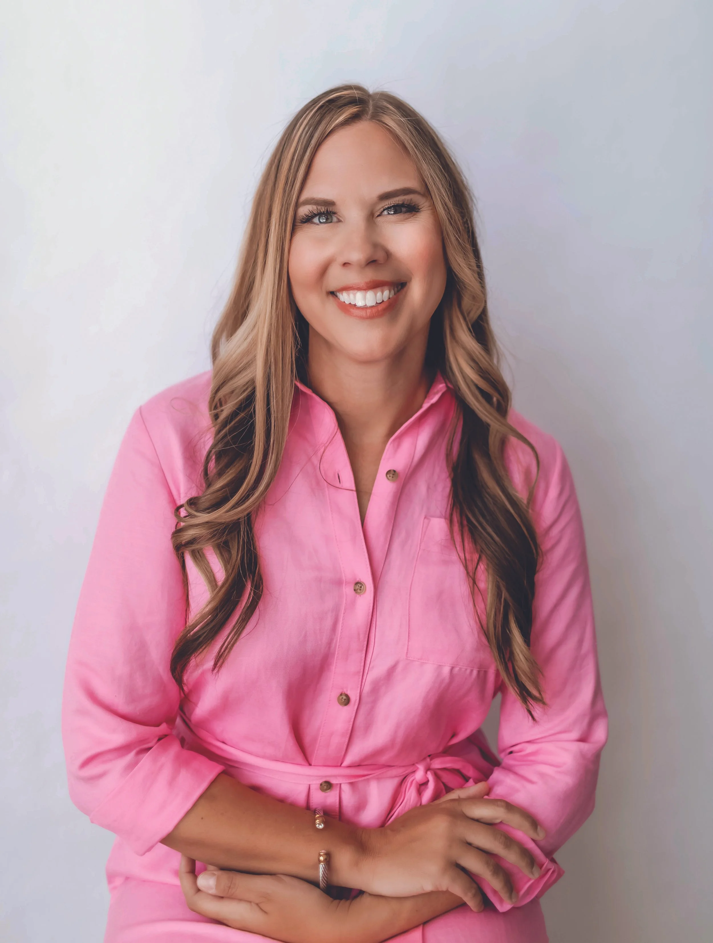 A woman with long wavy blonde hair wearing a pink button-up dress smiling with arms crossed against a plain white wall.