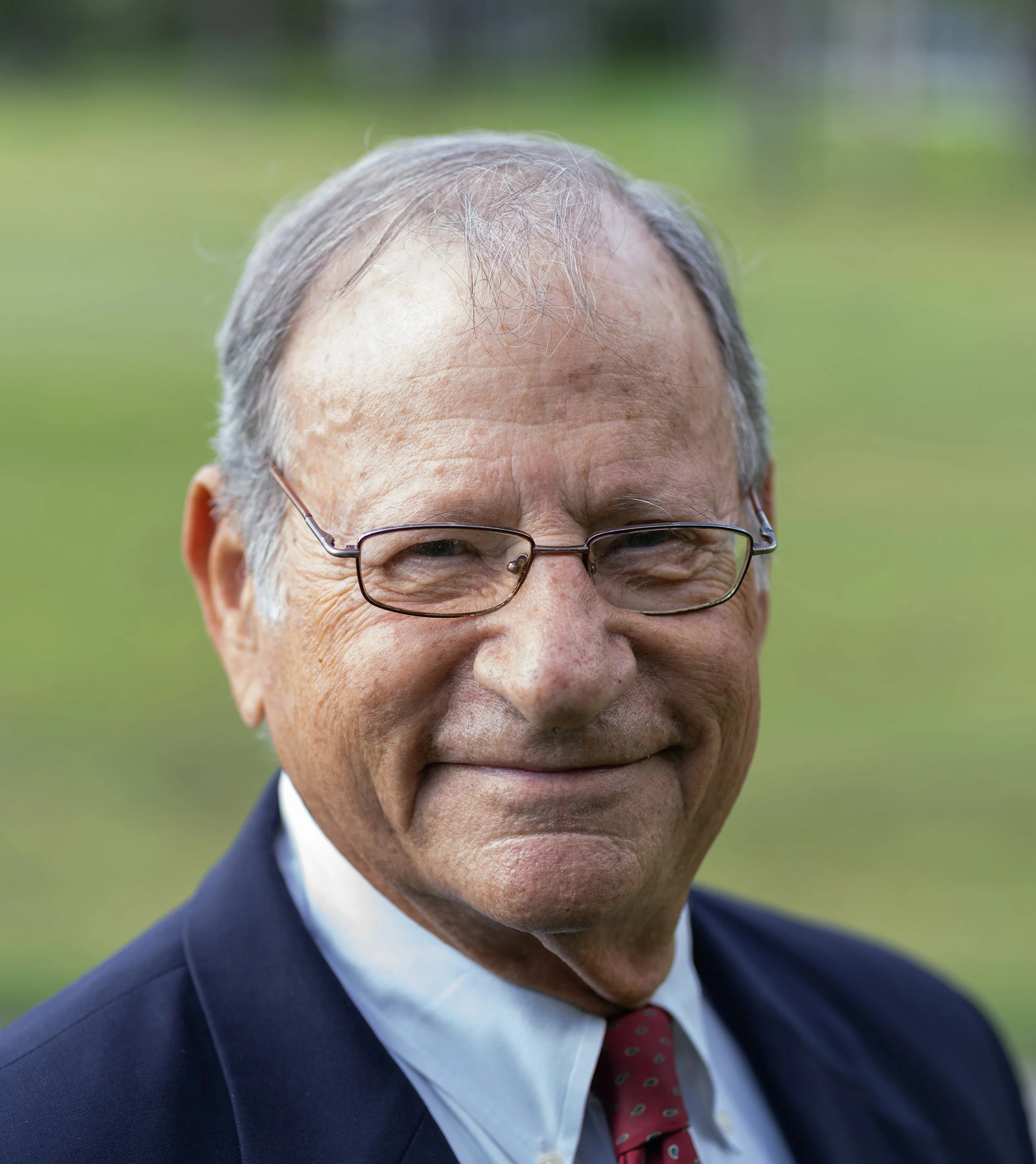 Close-up of an elderly man with glasses, smiling outdoors, wearing a suit and tie.