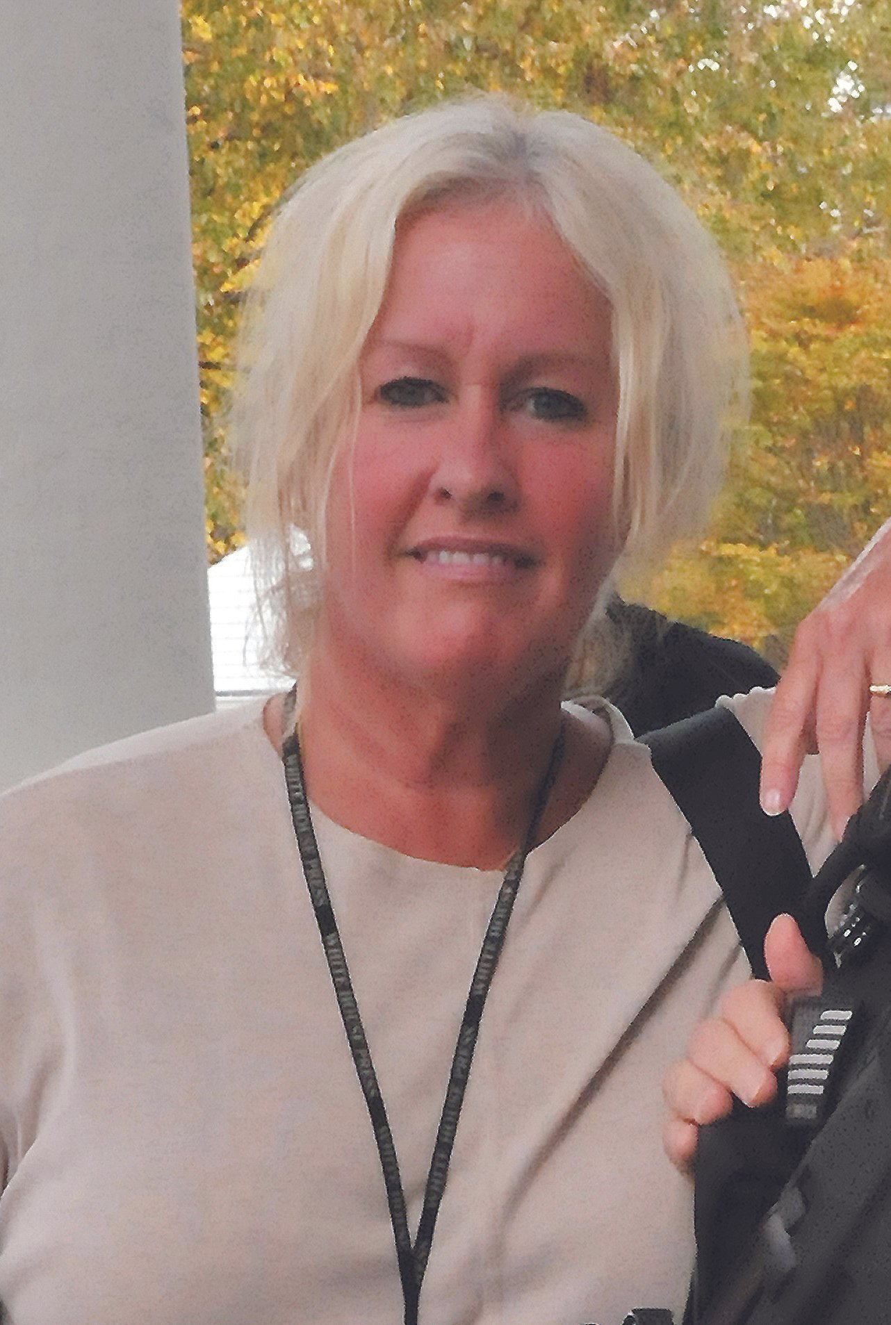 A middle-aged woman with short blonde hair smiling at the camera, wearing a light-colored top and a lanyard around her neck, standing outdoors with trees with fall foliage in the background.