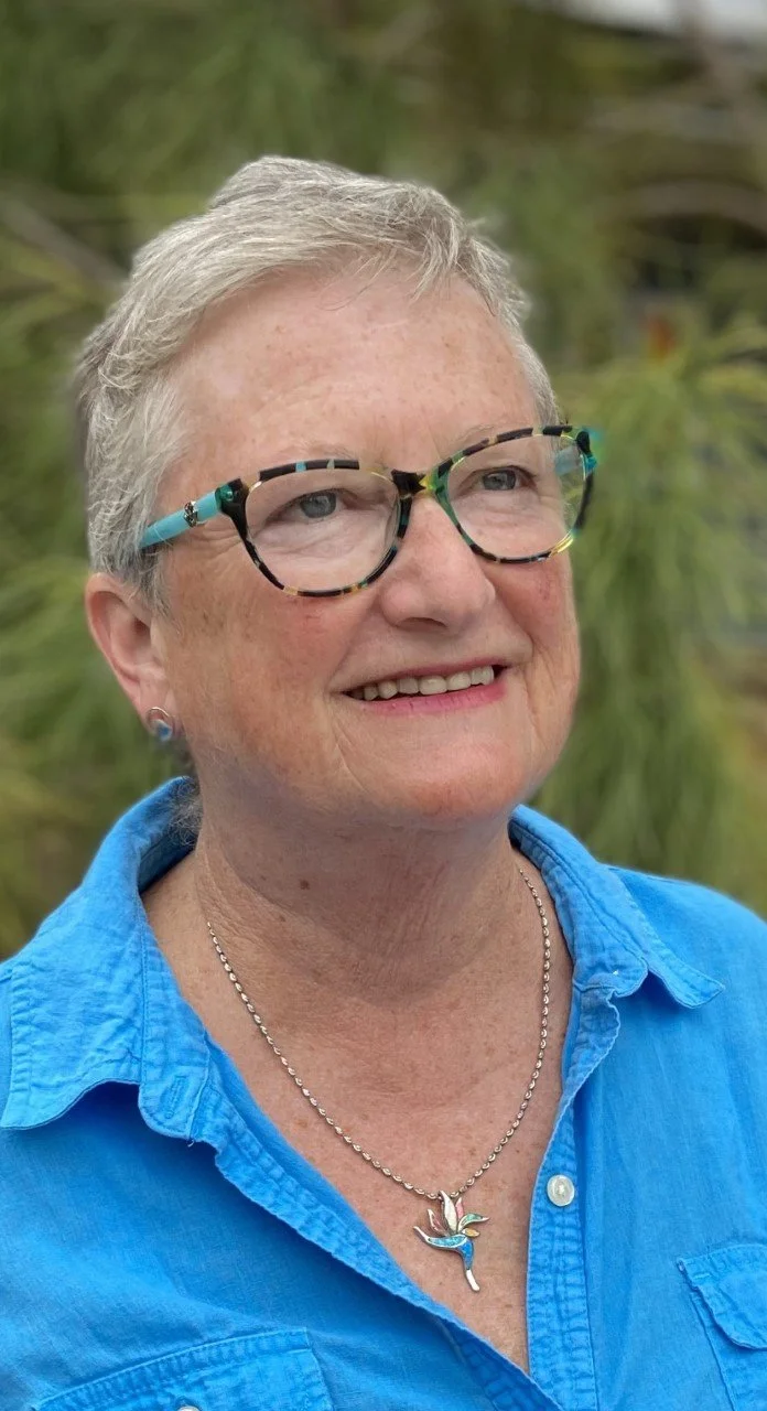 A smiling older woman with short gray hair, wearing large colorful glasses, a blue shirt, and a necklace with a hummingbird pendant, outdoors with green foliage in the background.