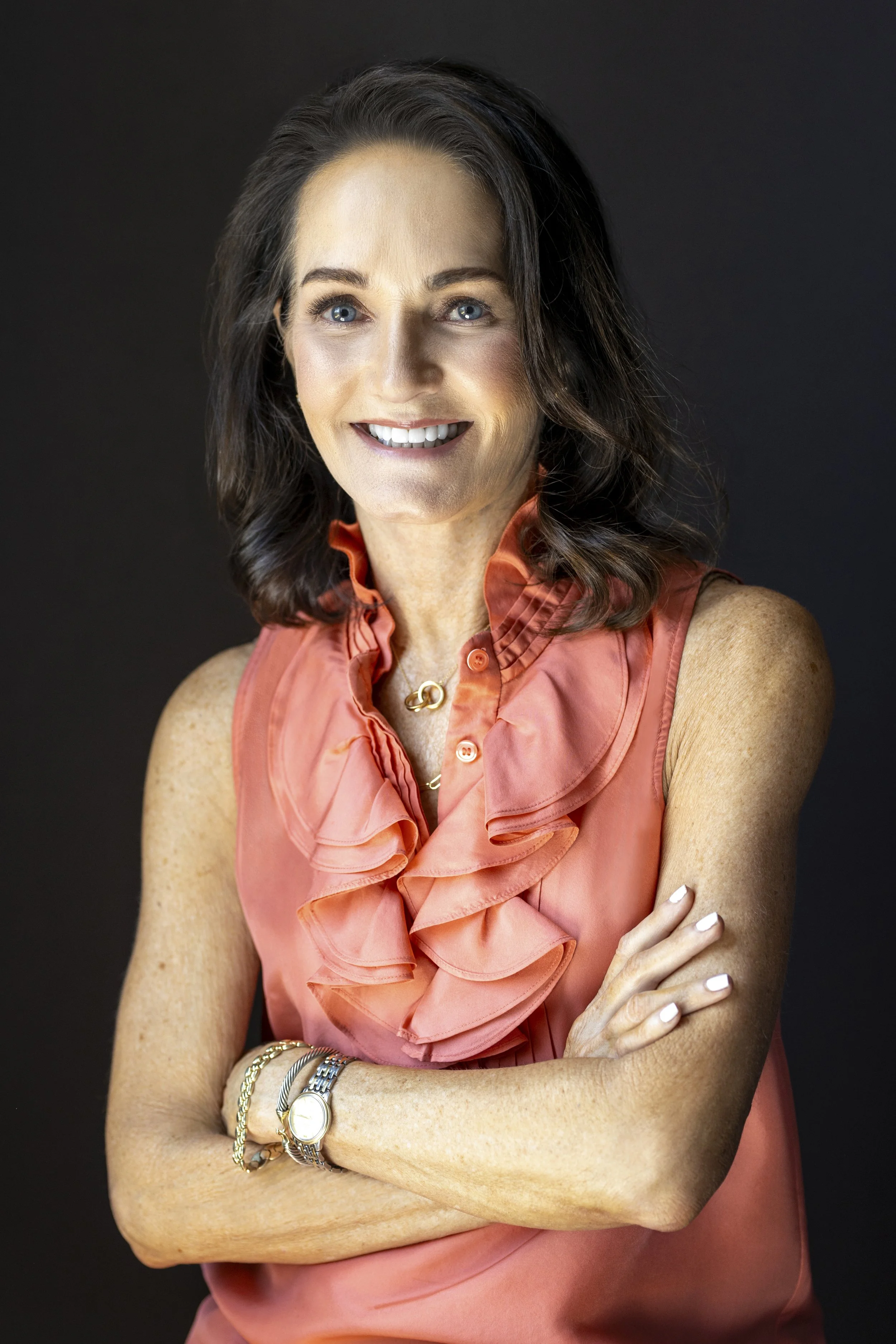A woman with dark brown hair, fair skin, and blue eyes smiling, wearing a coral sleeveless blouse with ruffles, a gold necklace, and a watch, standing against a dark background.