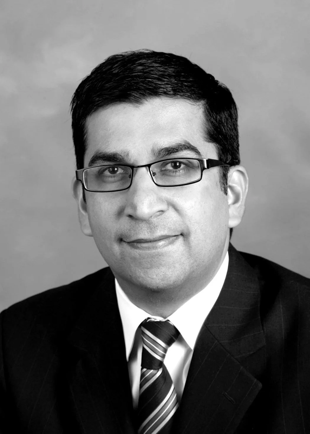 Black and white professional headshot of a man wearing glasses, a suit, and a striped tie, smiling softly.