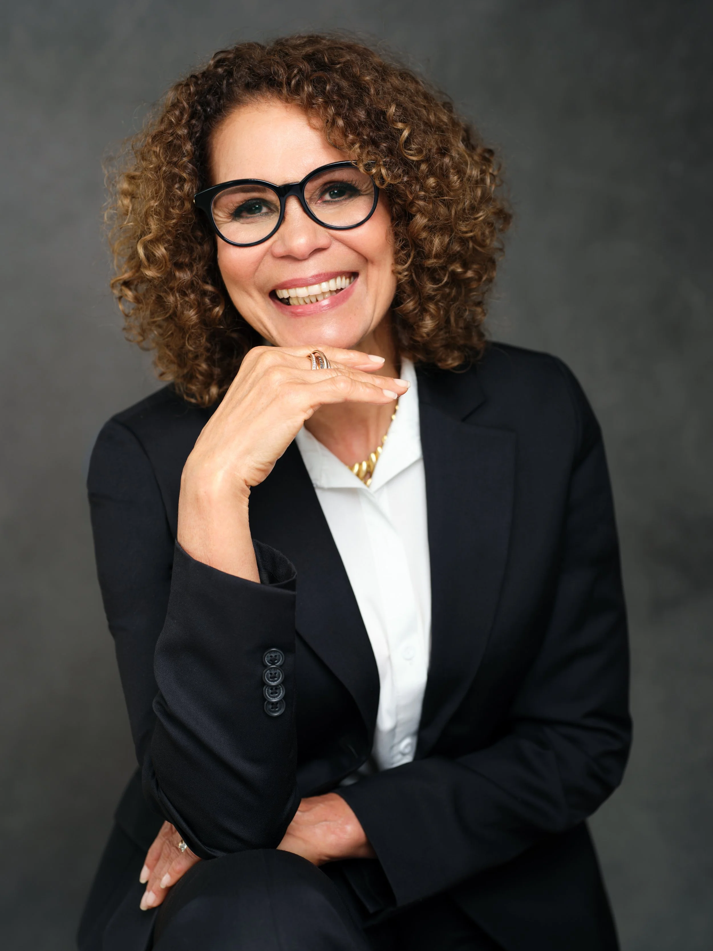 A woman with curly hair, wearing glasses, and a professional black blazer, smiling at the camera against a dark grey background.