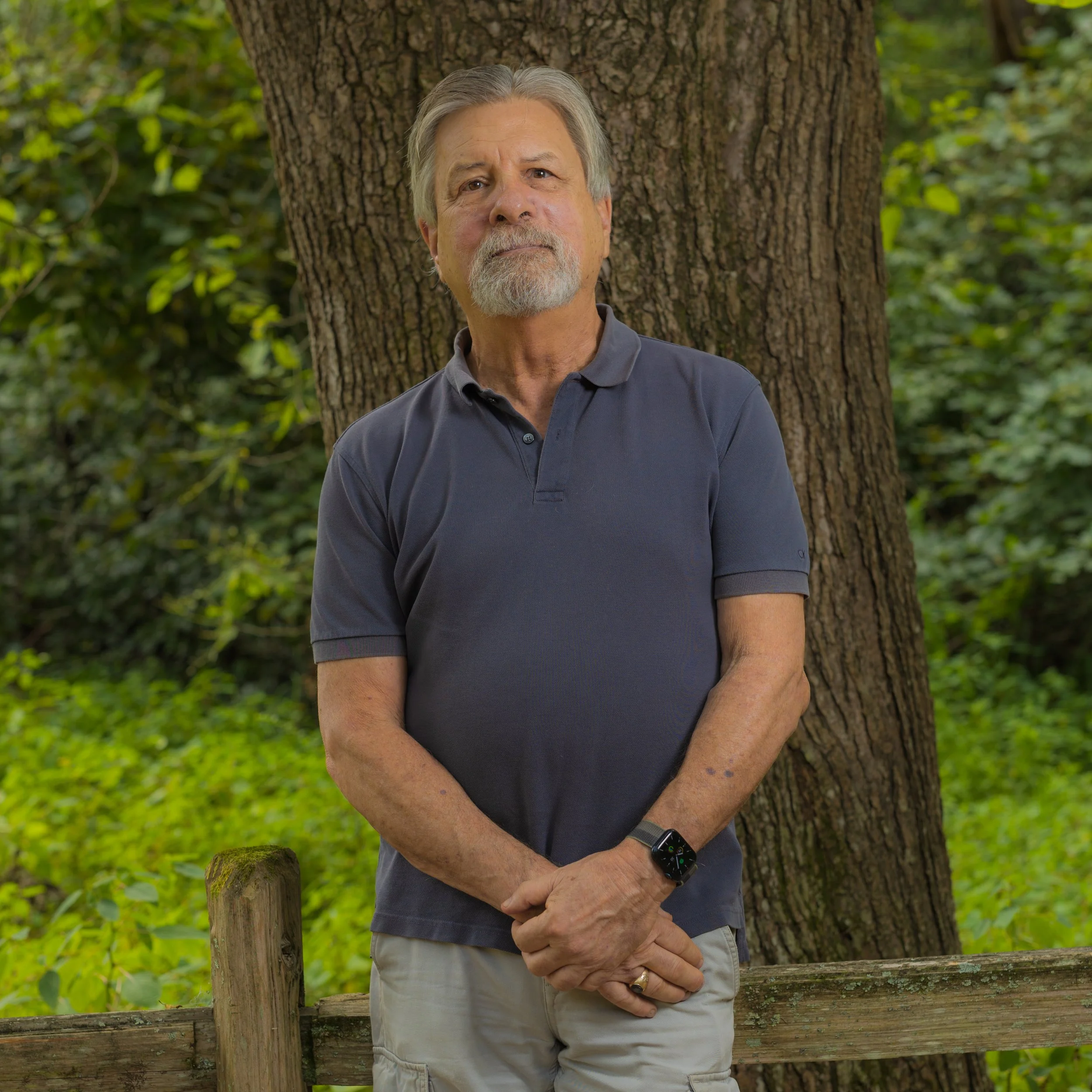 An older man with gray hair and beard standing outdoors near a tree, wearing a dark polo shirt, khaki shorts, and a smartwatch, with his hands clasped in front of him.