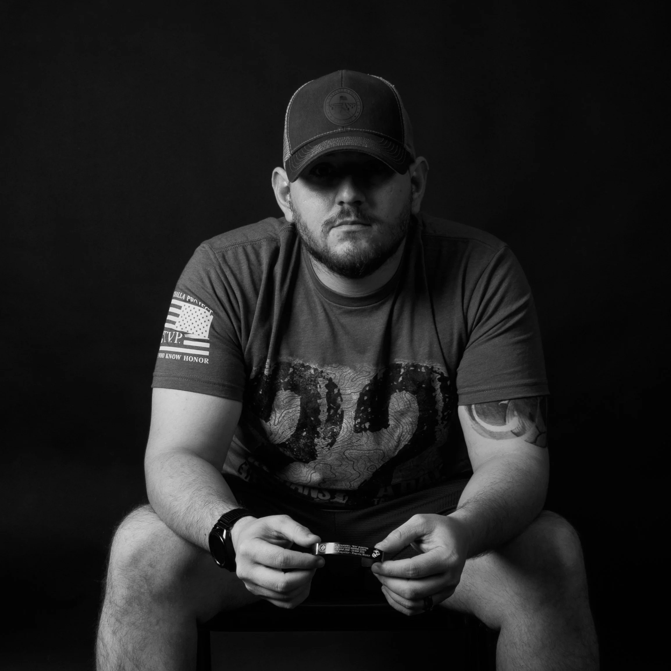 A man sitting on a chair, wearing a cap, t-shirt with a graphic design, and a watch, holding a small object in his hands, against a dark background.