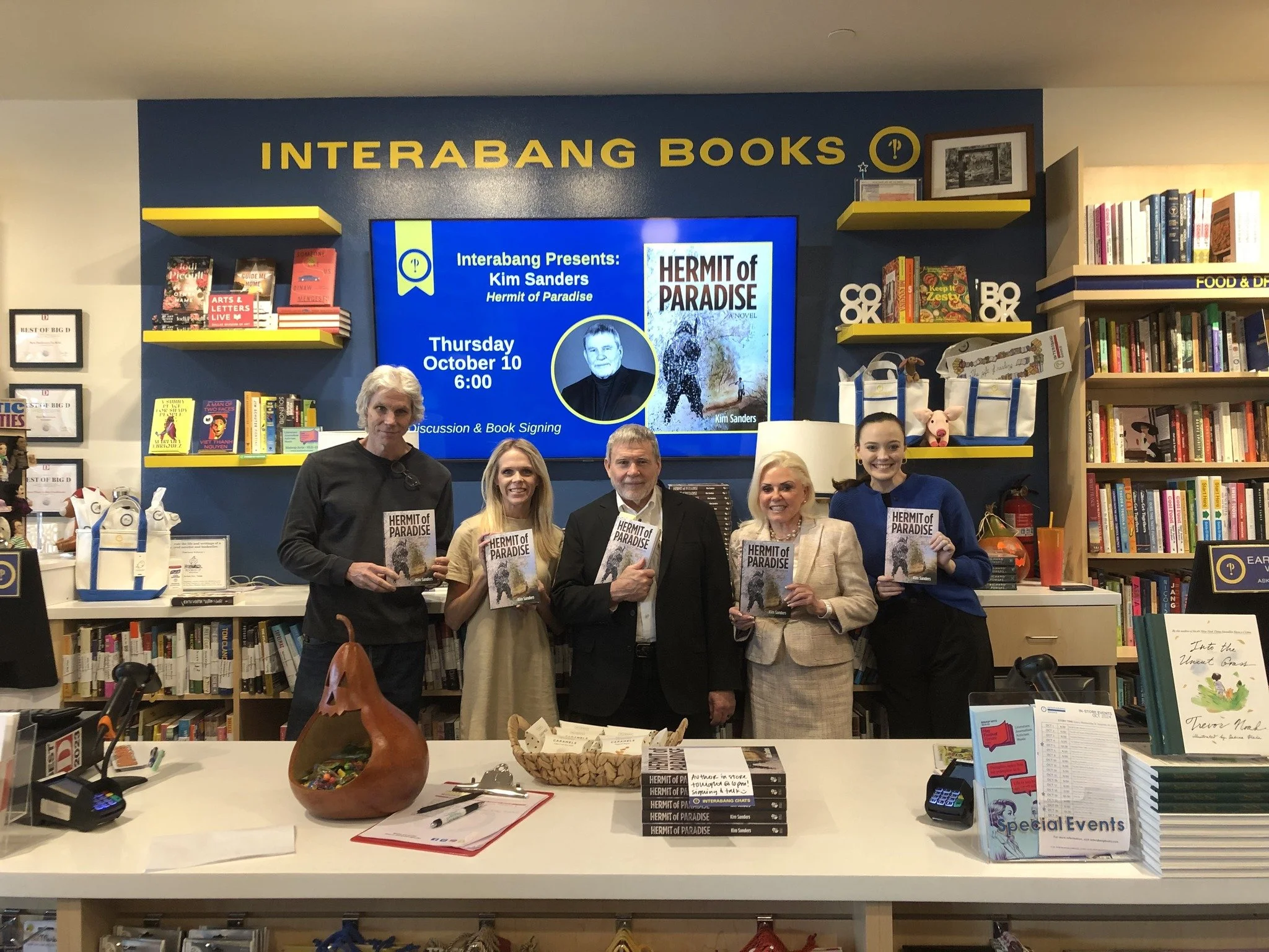 Group of six people holding copies of the book 'Hermit of Paradise' in a bookstore event. Bookshelves and promotional screen behind them showing the author's name Kim Sanders and the book cover.