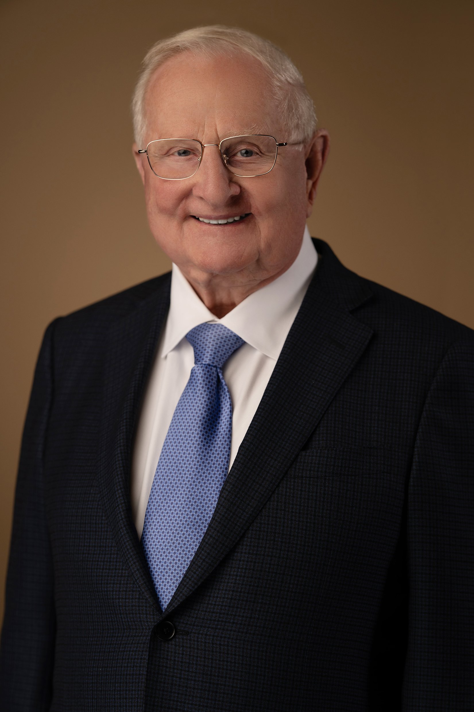 Portrait of an elderly man wearing glasses, a dark suit, white shirt, and a light blue tie, smiling against a plain background.