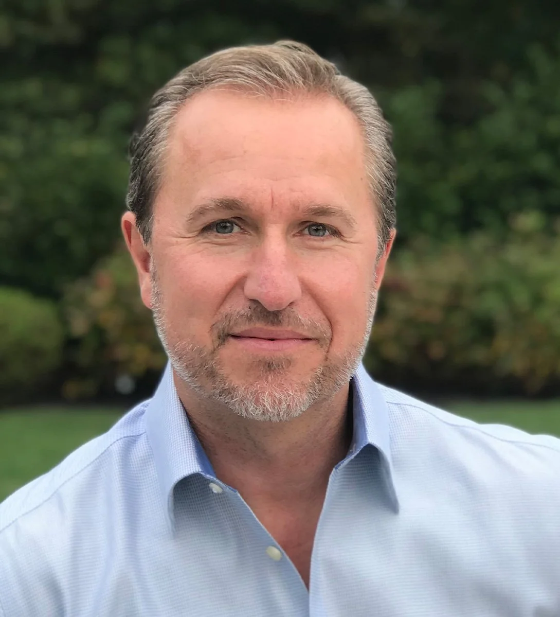 A middle-aged man with light brown hair, blue eyes, and a beard standing outdoors. He is wearing a light blue, collared shirt and is smiling slightly. The background features greenery with trees and grass.