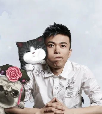 A man with short black hair wearing a white shirt sitting at a table with a surprised expression. To his left, there's a dog wearing glasses and a flower crown, and to his right, a black and white cat plush and a large stuffed cat behind him.
